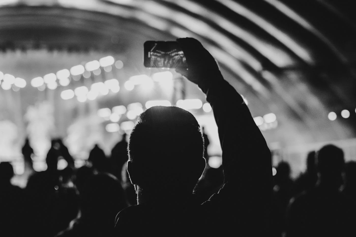 Silhouette of a person holding a smartphone up in a crowd at a concert or event with bright stage lights in the background.