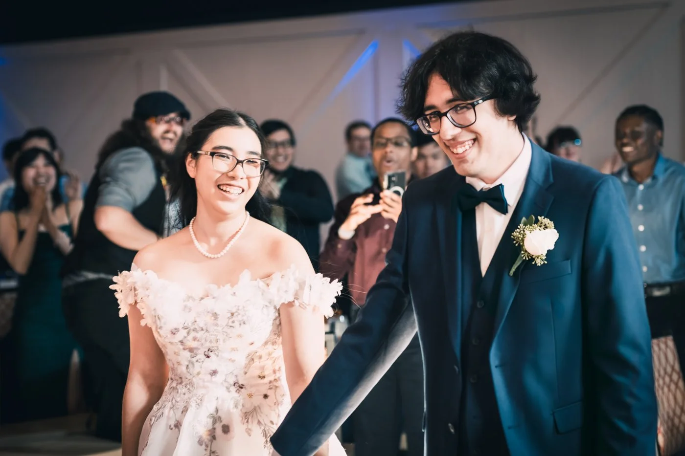 A bride and groom smiling at each other during their wedding reception, surrounded by guests, with one guest taking a photo in the background.