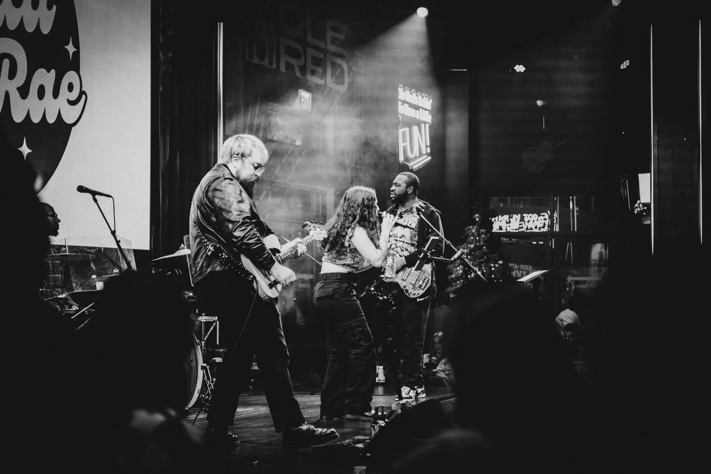 Musicians performing on stage in a black and white photo, with a female singer with curly hair singing into a microphone. The band includes a guitarist with long hair, a bass player, and a drummer slightly visible in the background, in a venue with neon signs and a large screen.