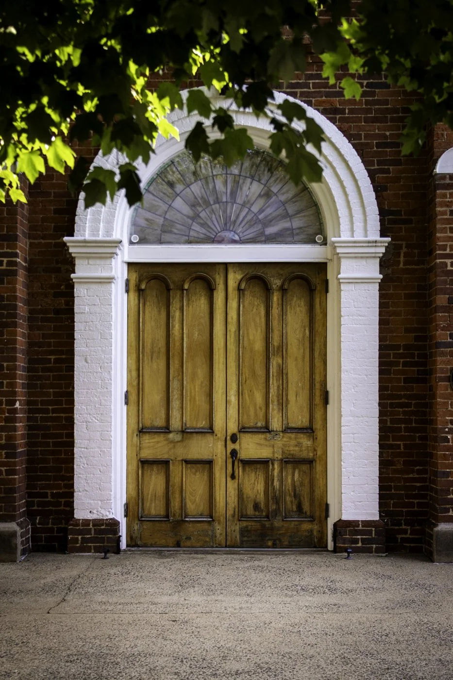 Wooden double doors set in a brick building, with a semi-circular stained glass window above and white decorative trim around the entrance, partially obscured by green tree leaves.