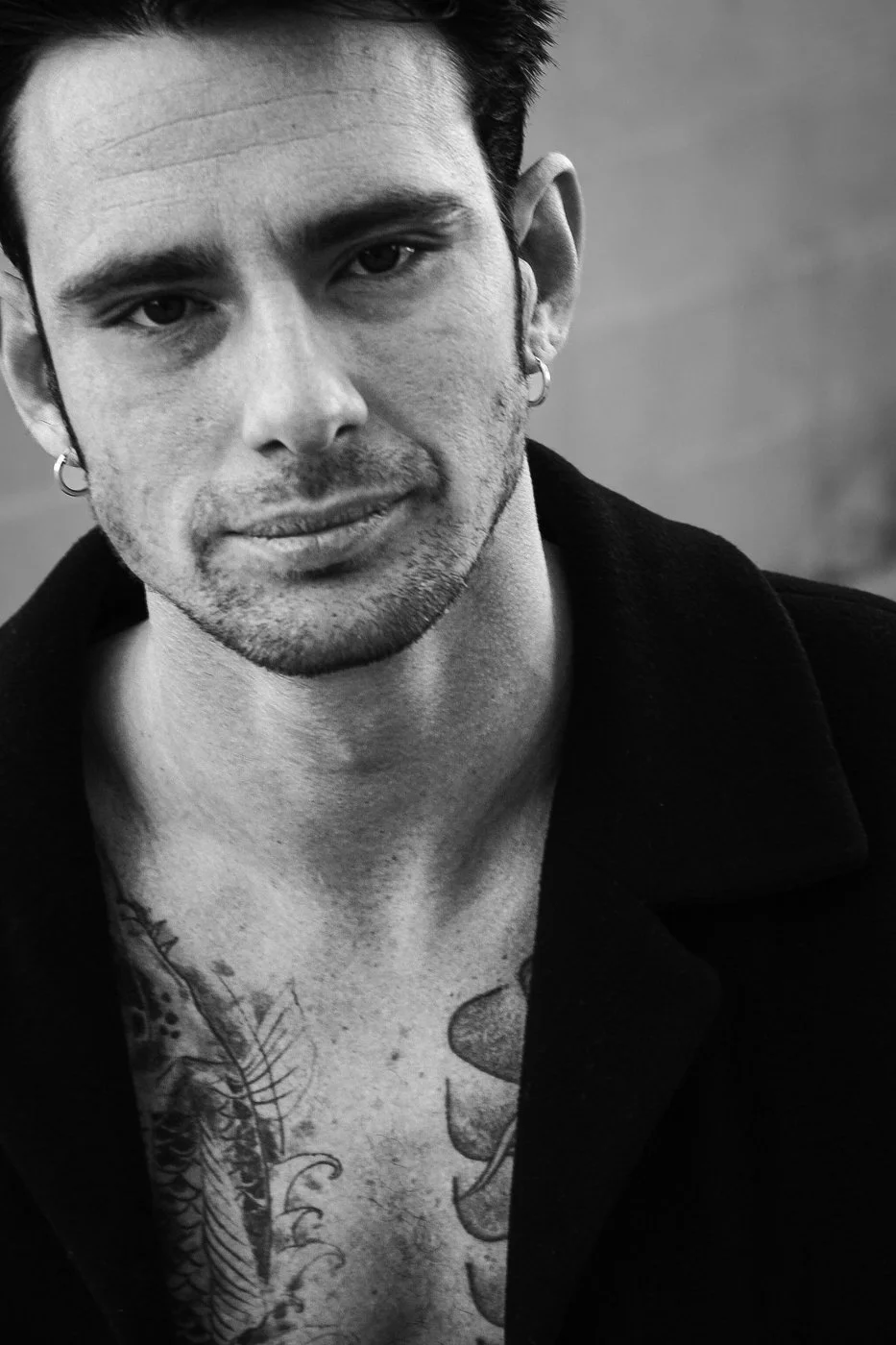 A black and white portrait of a young man with short dark hair, earrings, and visible tattoos on his chest. He has a slight smile and is wearing a dark jacket.