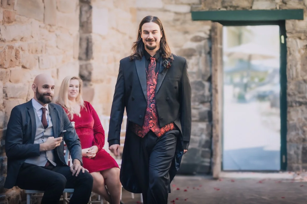 A man with long hair and a beard walking down the aisle at a wedding ceremony, with two seated guests, a man and a woman, watching and smiling inside a rustic stone building with an open door showing outdoor scenery.