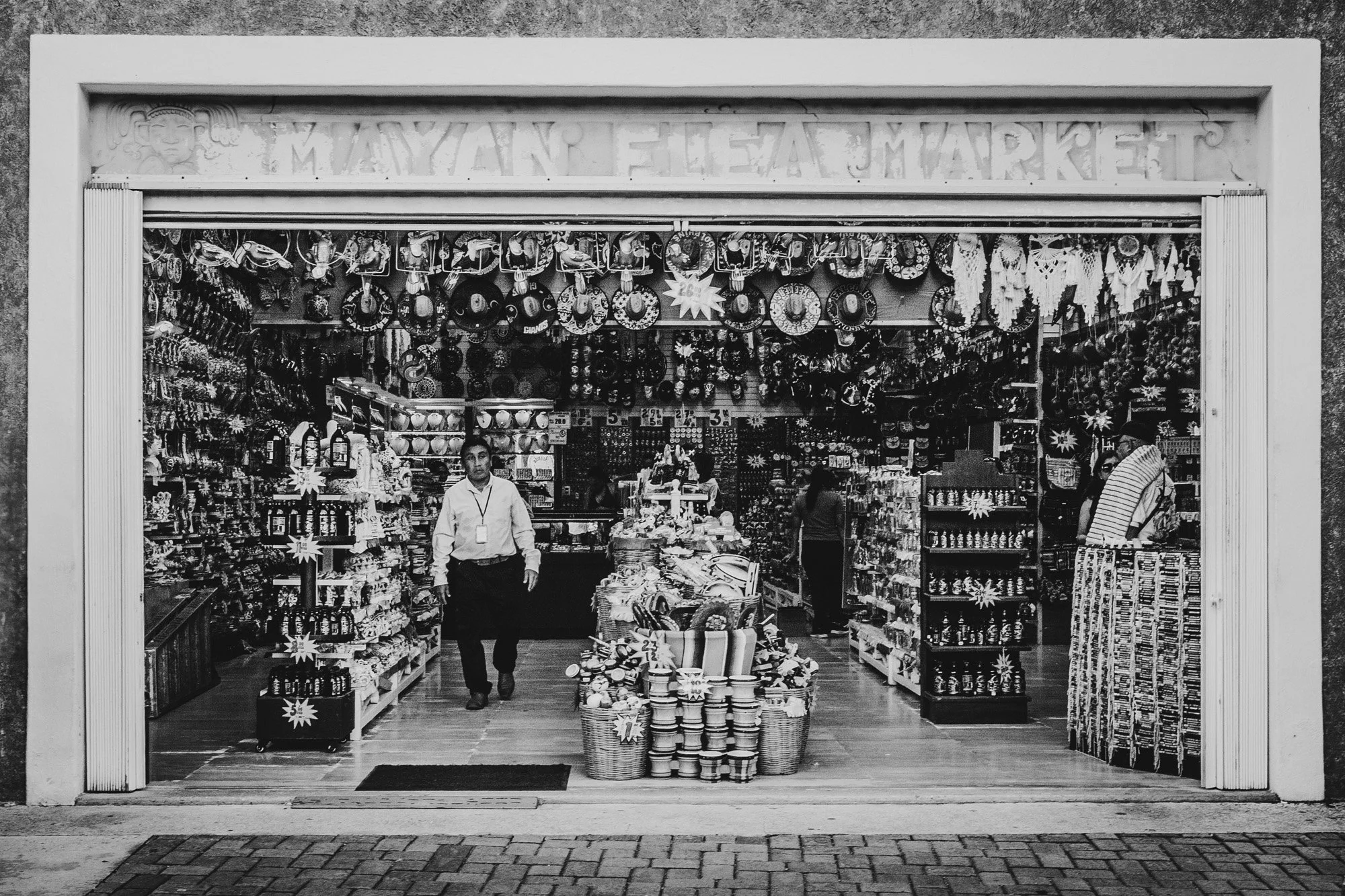 Black and white photo of a store entrance with the sign 'MAYAN FLORA MARKET' above, showcasing shelves filled with various souvenirs, hats, and decorative items, and three people inside.