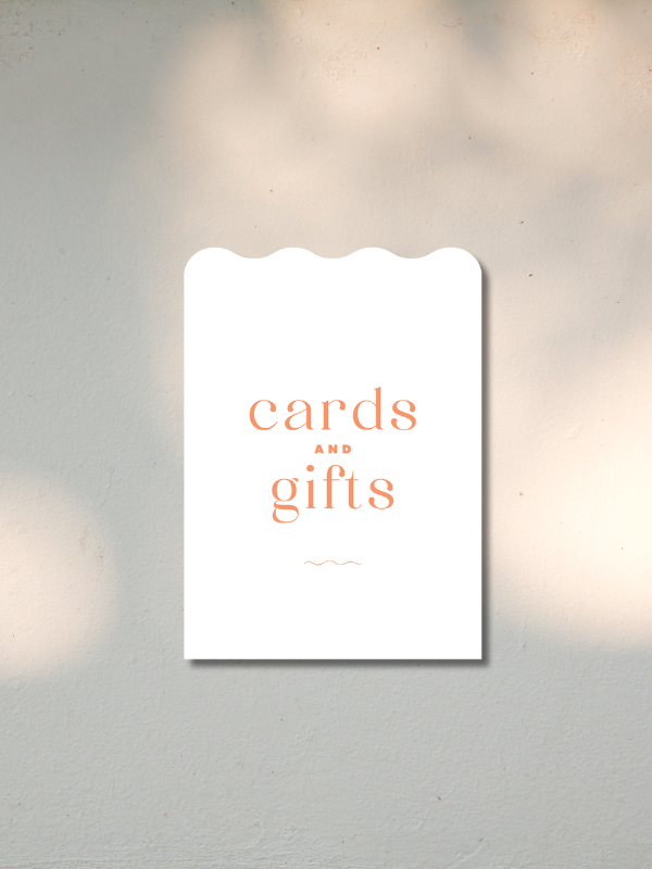 A white sign with scalloped top edge on a wall, reading "cards and gifts" in orange text.