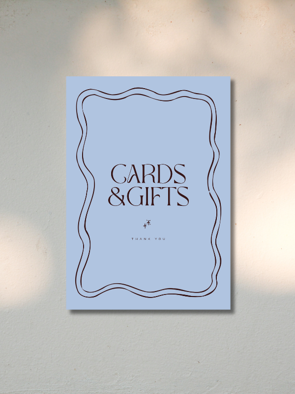 A light blue sign with a black decorative border that reads "Cards & Gifts" and "Thank You" beneath it, hanging on a white wall.