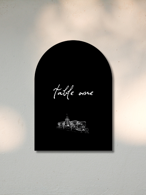 A black menu sign on a white wall with the words 'table one' written in elegant cursive and a white line drawing of a building at the bottom.