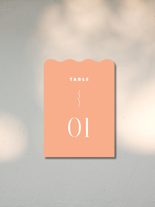 A peach-colored sign with the word 'TABLE' at the top, a wavy line in the middle, and the number '01' at the bottom, mounted on a light wall.
