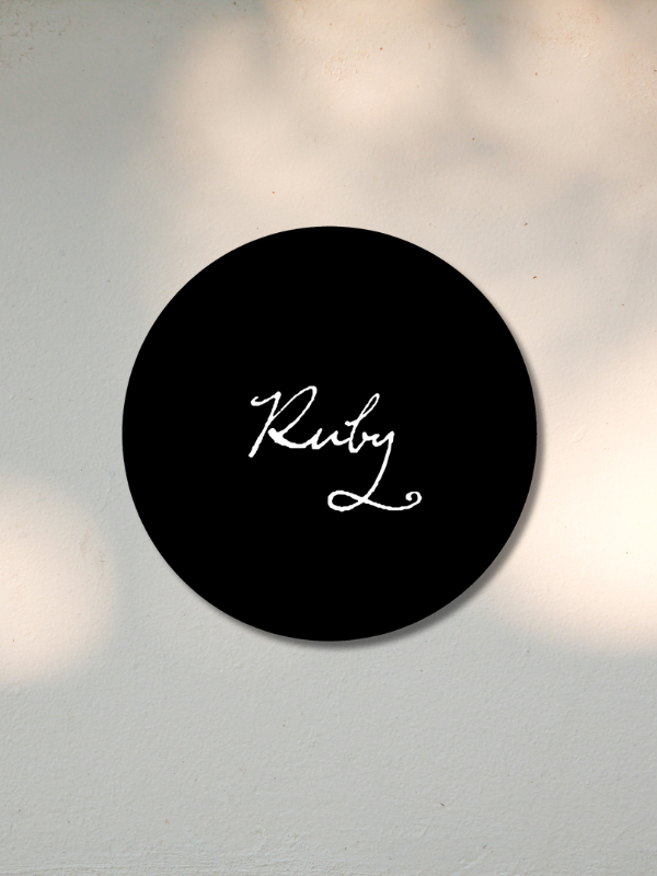 Black circular sign with the word 'Ruby' written in white cursive font on a light-colored wall.