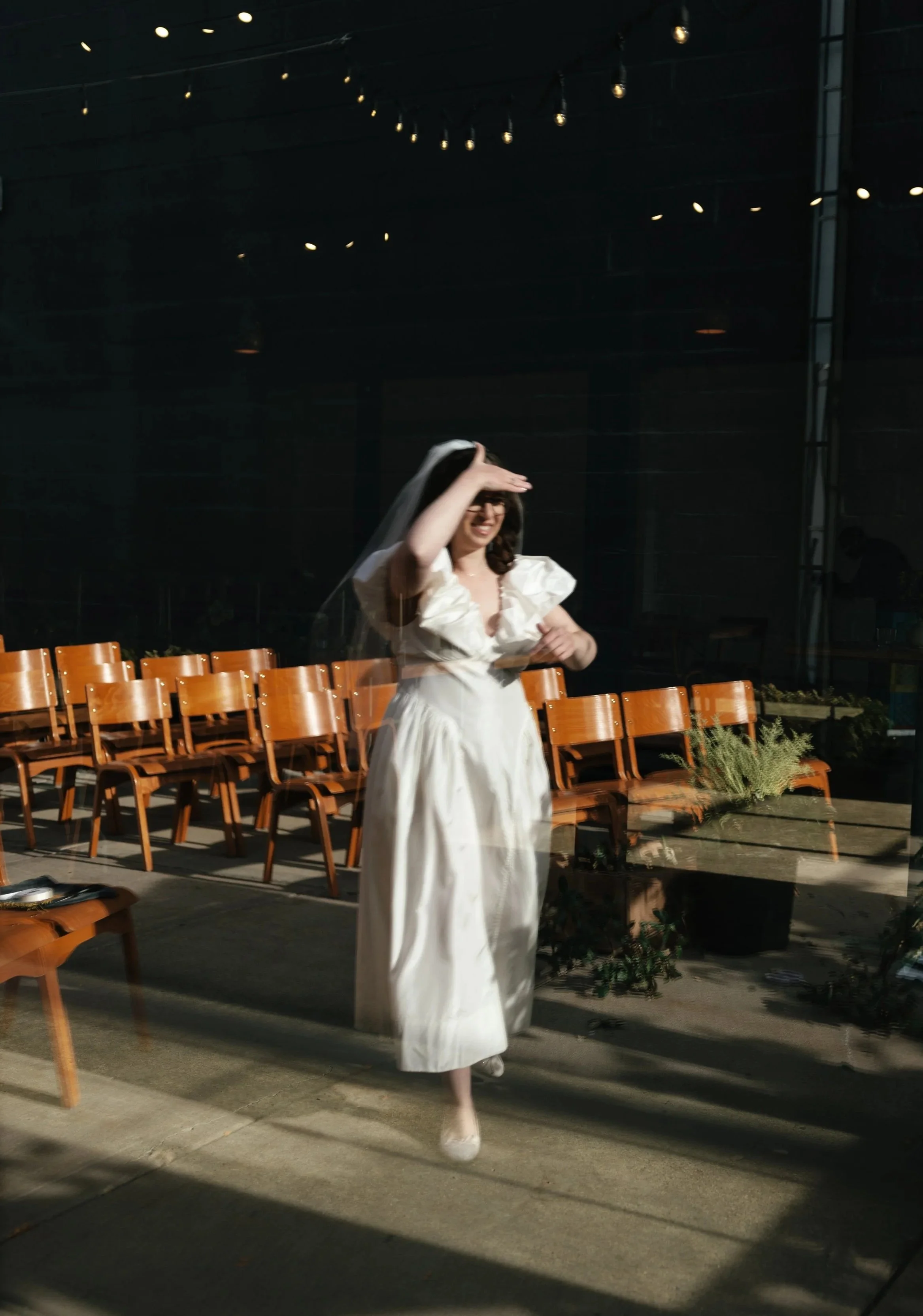 A bride in a white dress and veil is smiling and standing among empty wooden chairs in a dimly lit St. Paul Minnesota venue with string lights overhead, outdoors or in a spacious area.