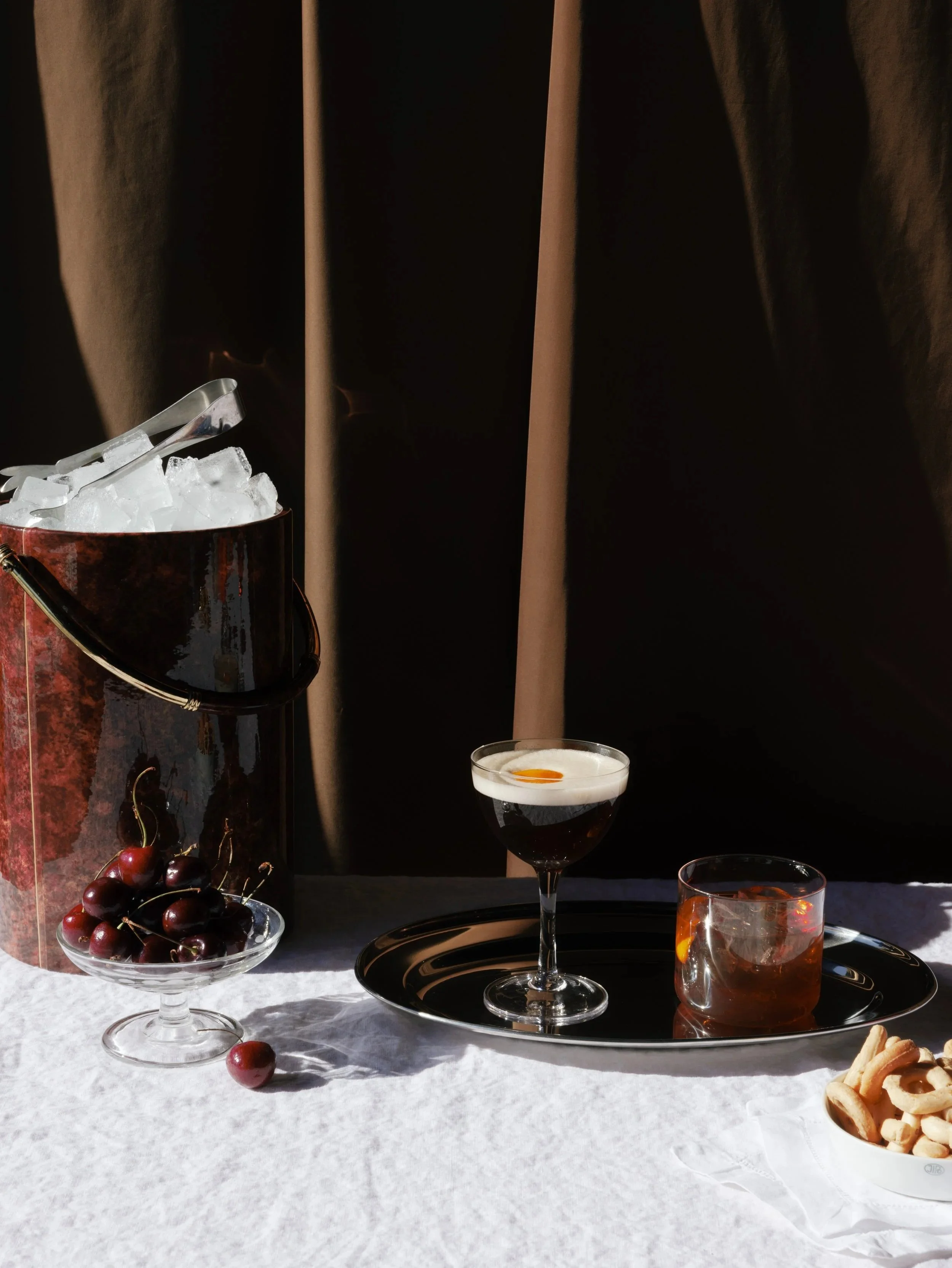 A table set with cocktails, cherries, a bowl of cookies, and a ice bucket with tongs, against a dark curtain backdrop.