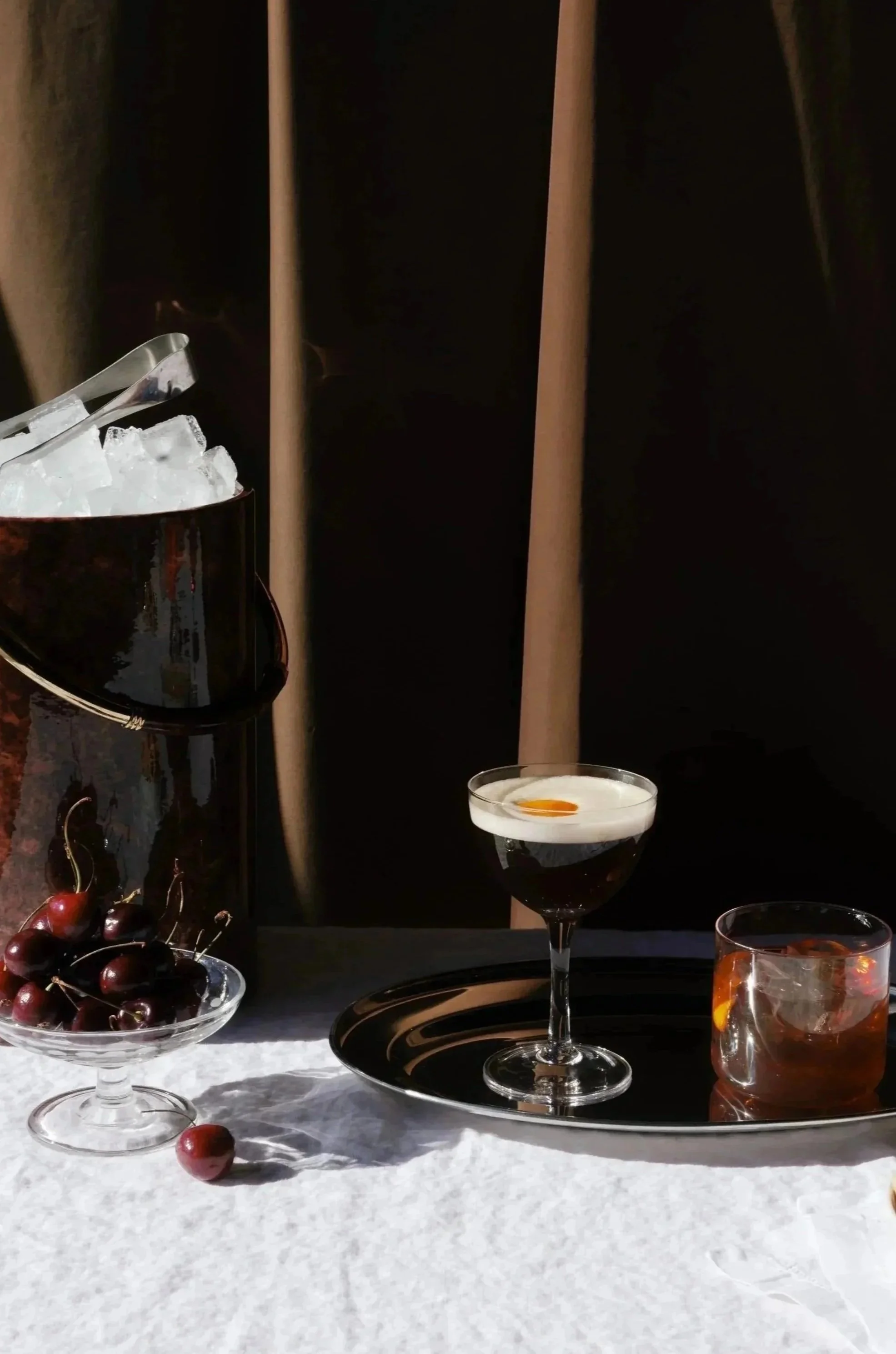 An espresso martini, a bowl of cherries, and old fashioned, and a bowl of ice cubes with tongs on a white surface, against a dark background.