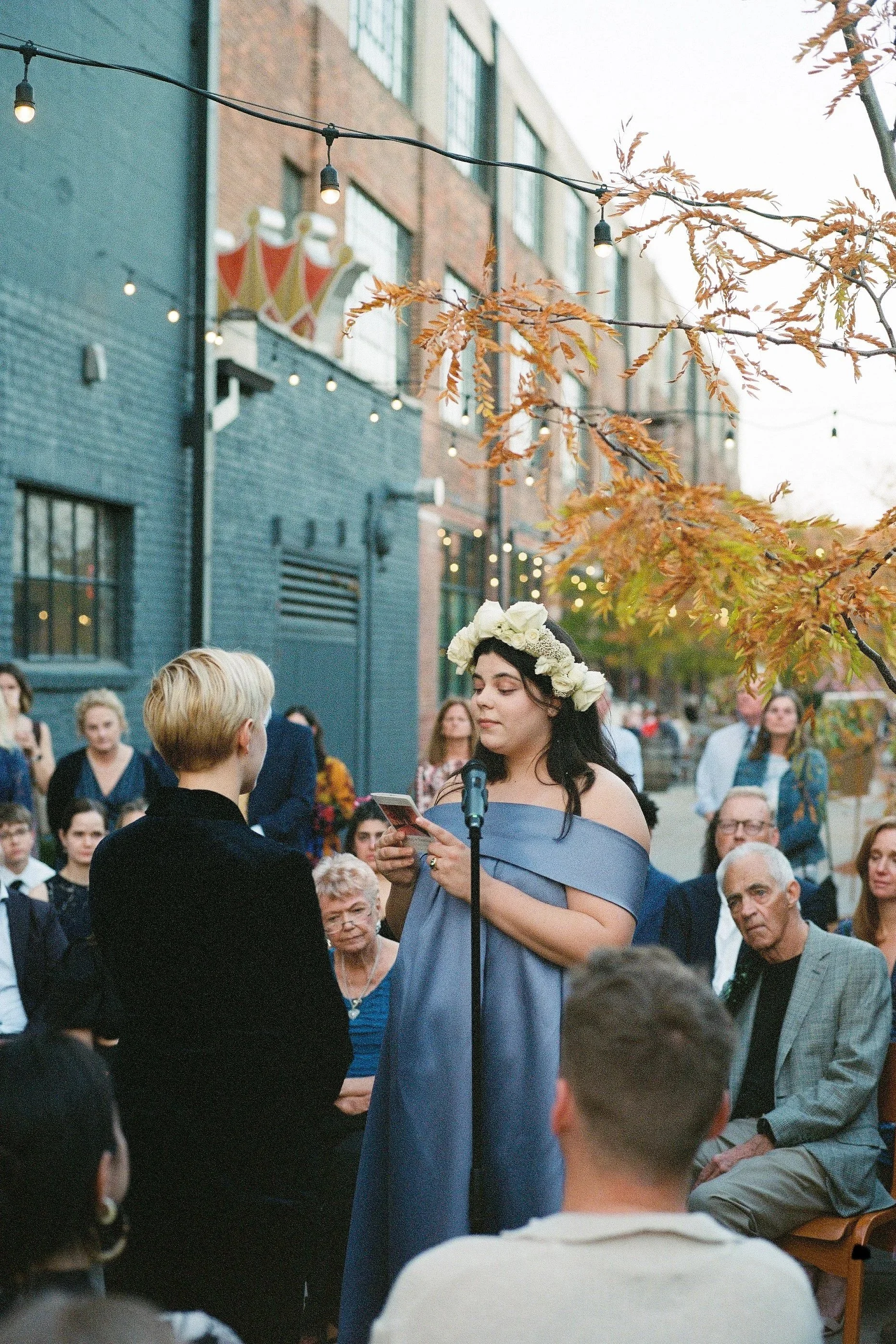 A woman with dark hair wearing a light blue off-shoulder dress and floral crown is speaking into a microphone at an outdoor event with a crowd of people in the background, during autumn.