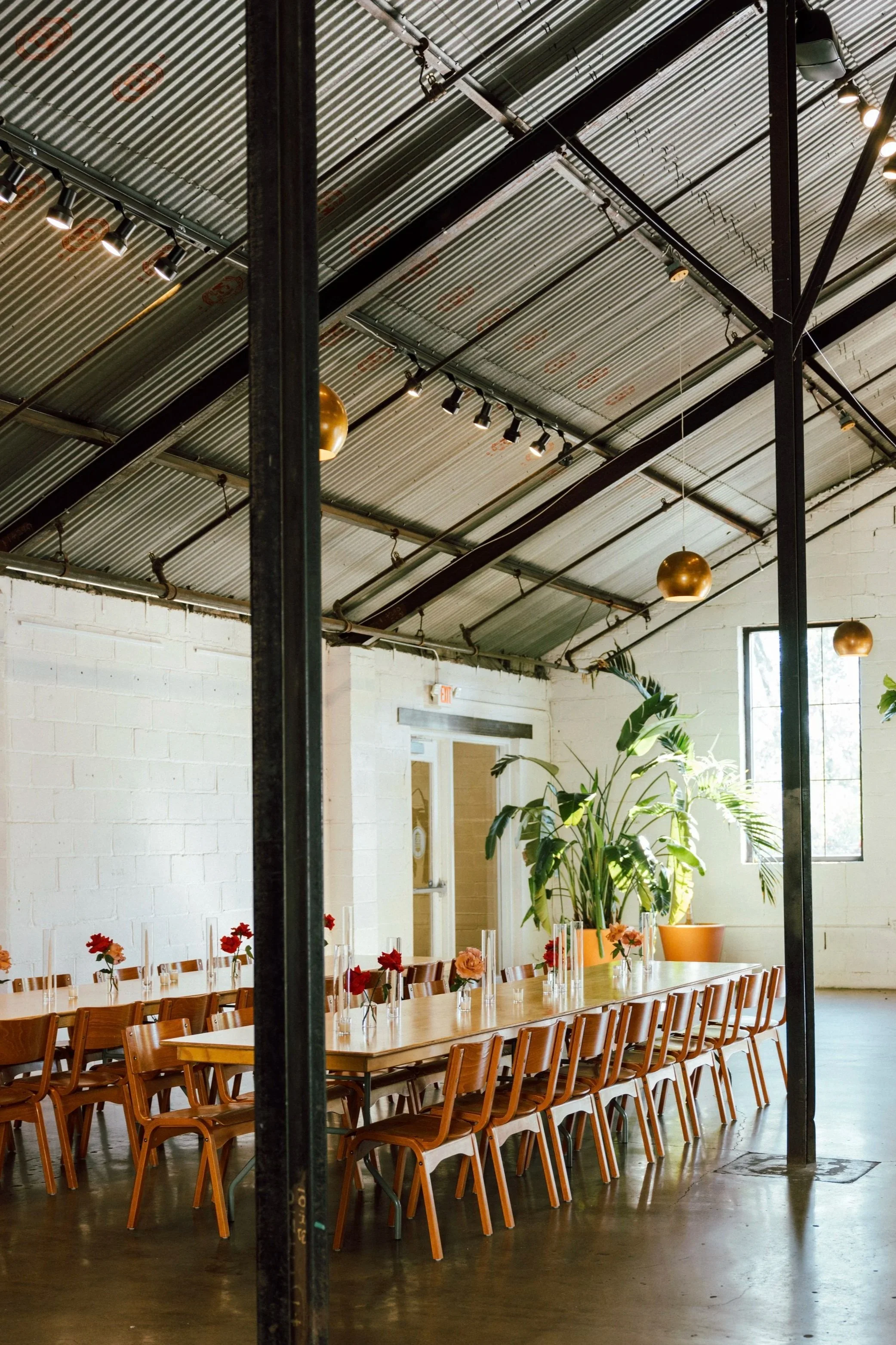 A large indoor space with a long wooden table set with small flower arrangements in glass vases, wooden chairs around the table, large green potted plants, industrial-style metal ceiling with hanging lights, and windows allowing natural light