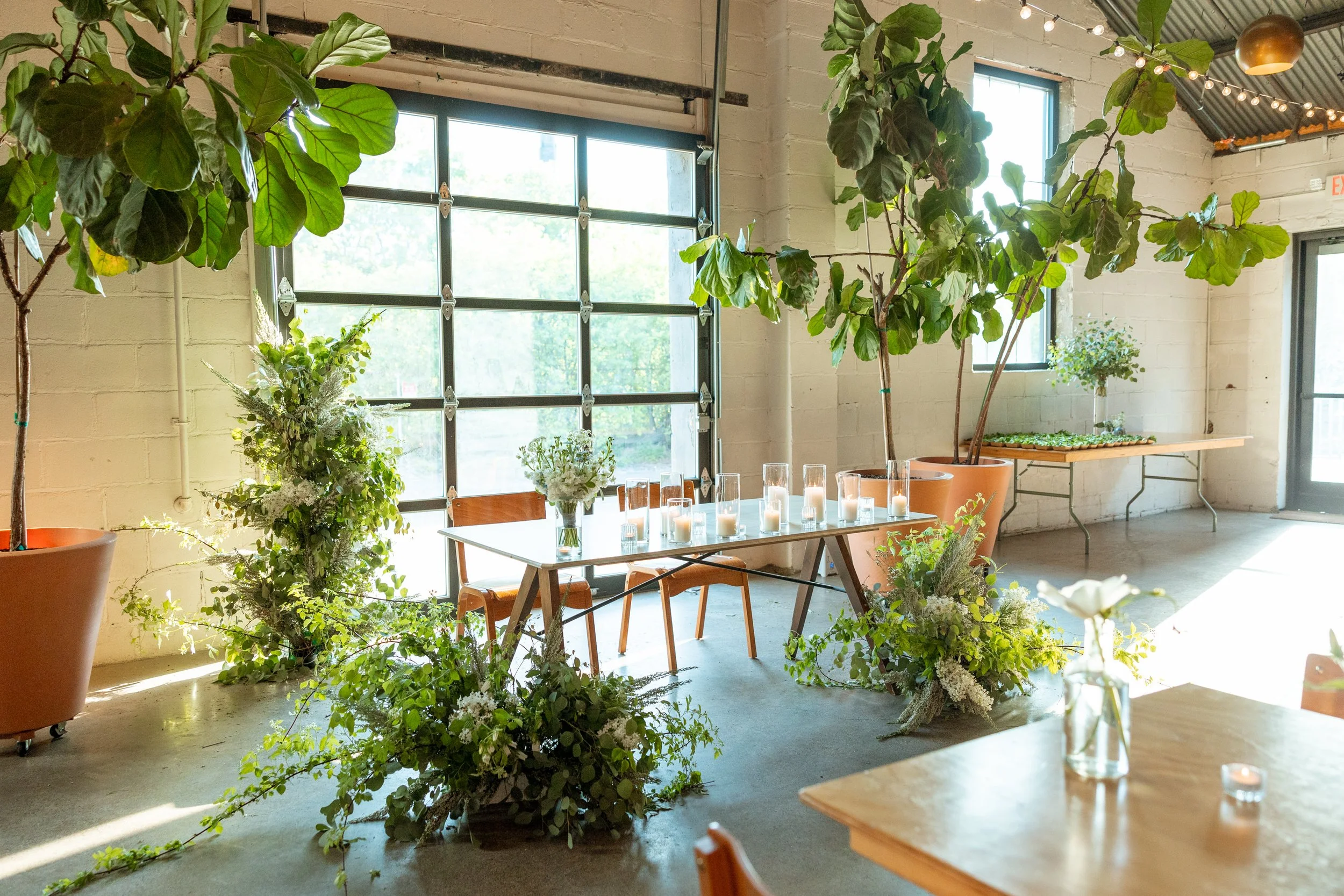 A white table with two chairs surrounded by green and white flowers on the floor and large potted plants behind