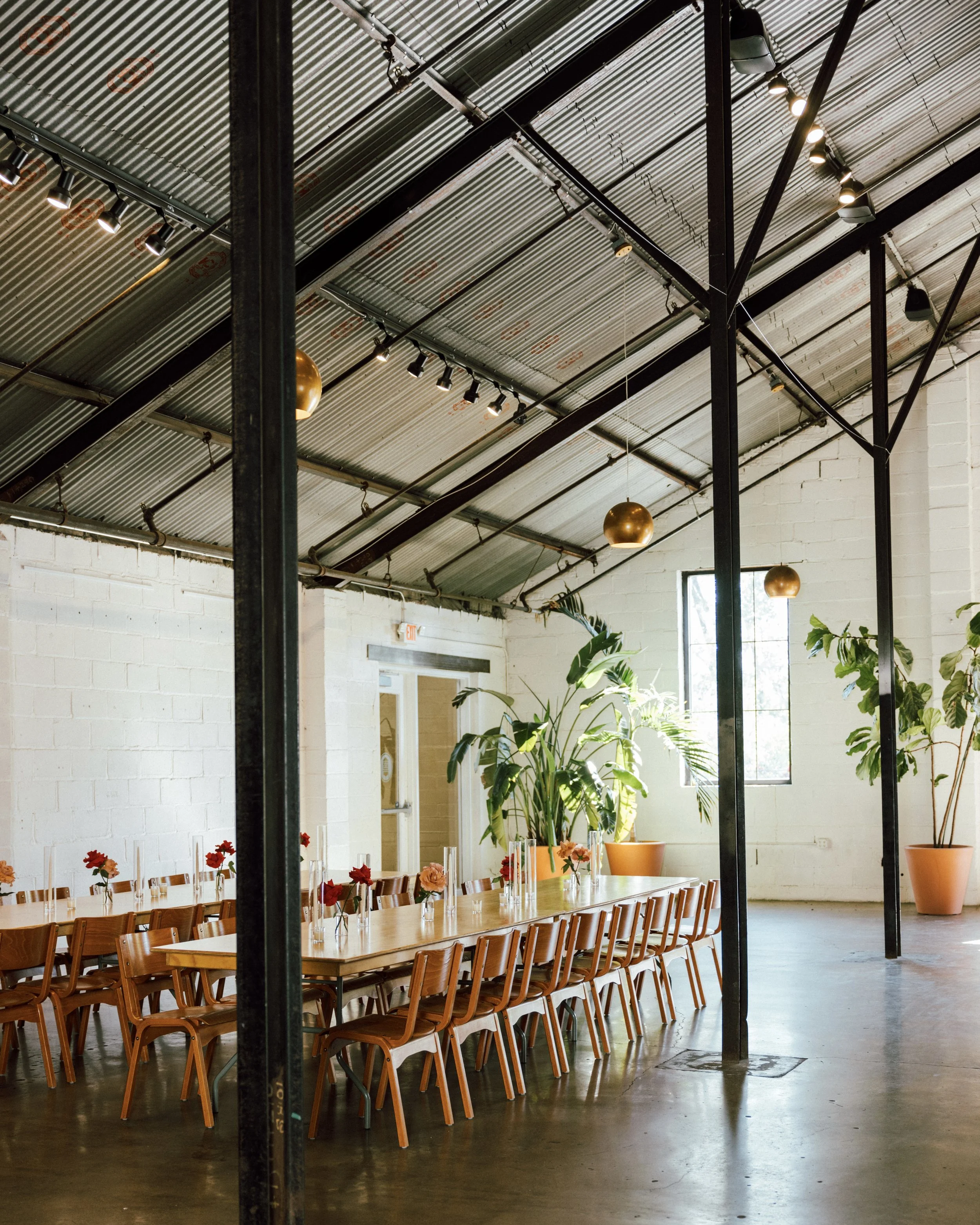 Long wooden dining table with chairs, decorated with small flower arrangements in glass vases, in a bright industrial-style room with large window, green plants in large pots, white brick walls, and a corrugated metal ceiling with track lighting.