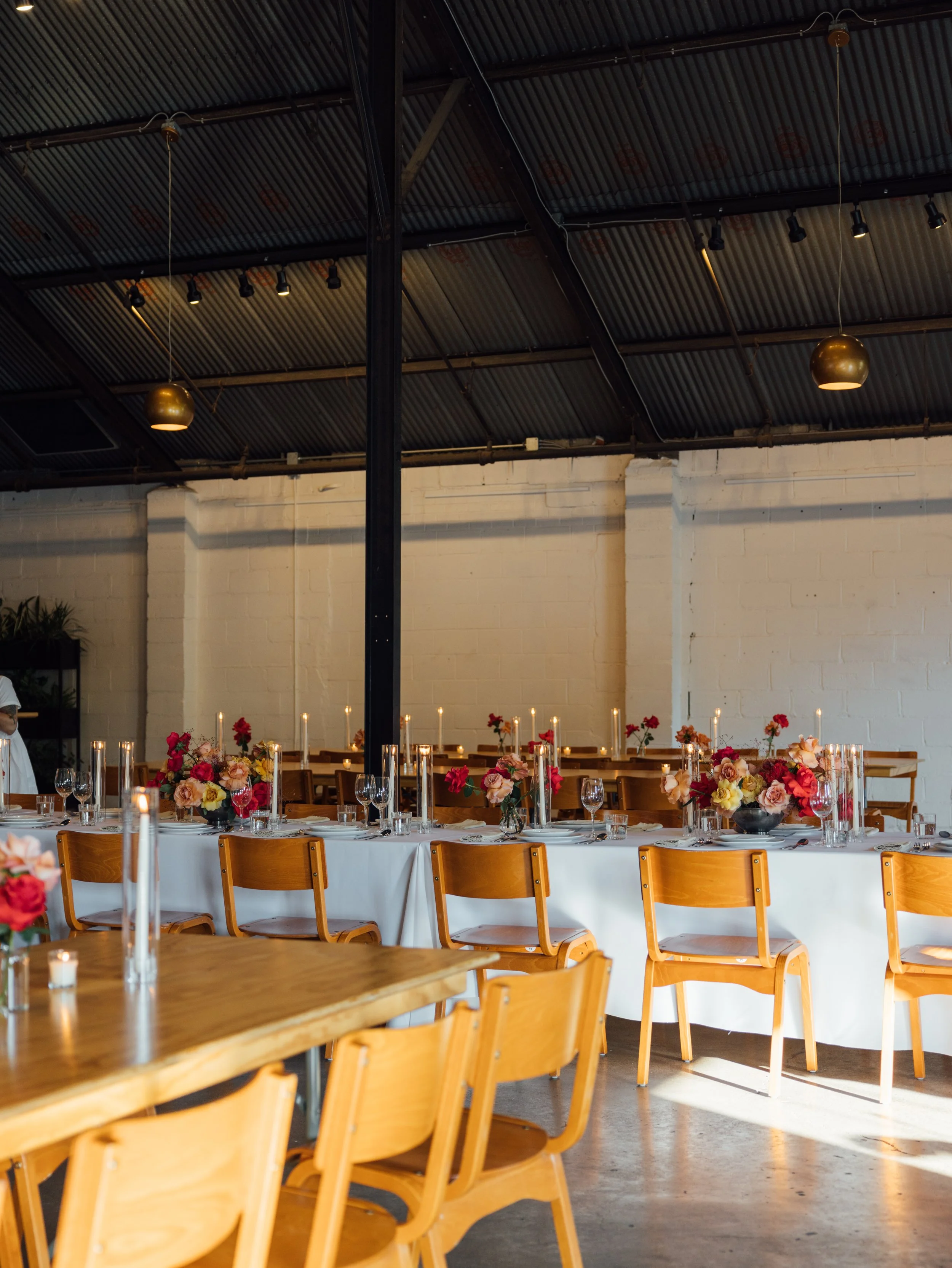 Event space decorated with a long white table with floral arrangements and candles, surrounded by wooden chairs, and a black metal ceiling with hanging lights.