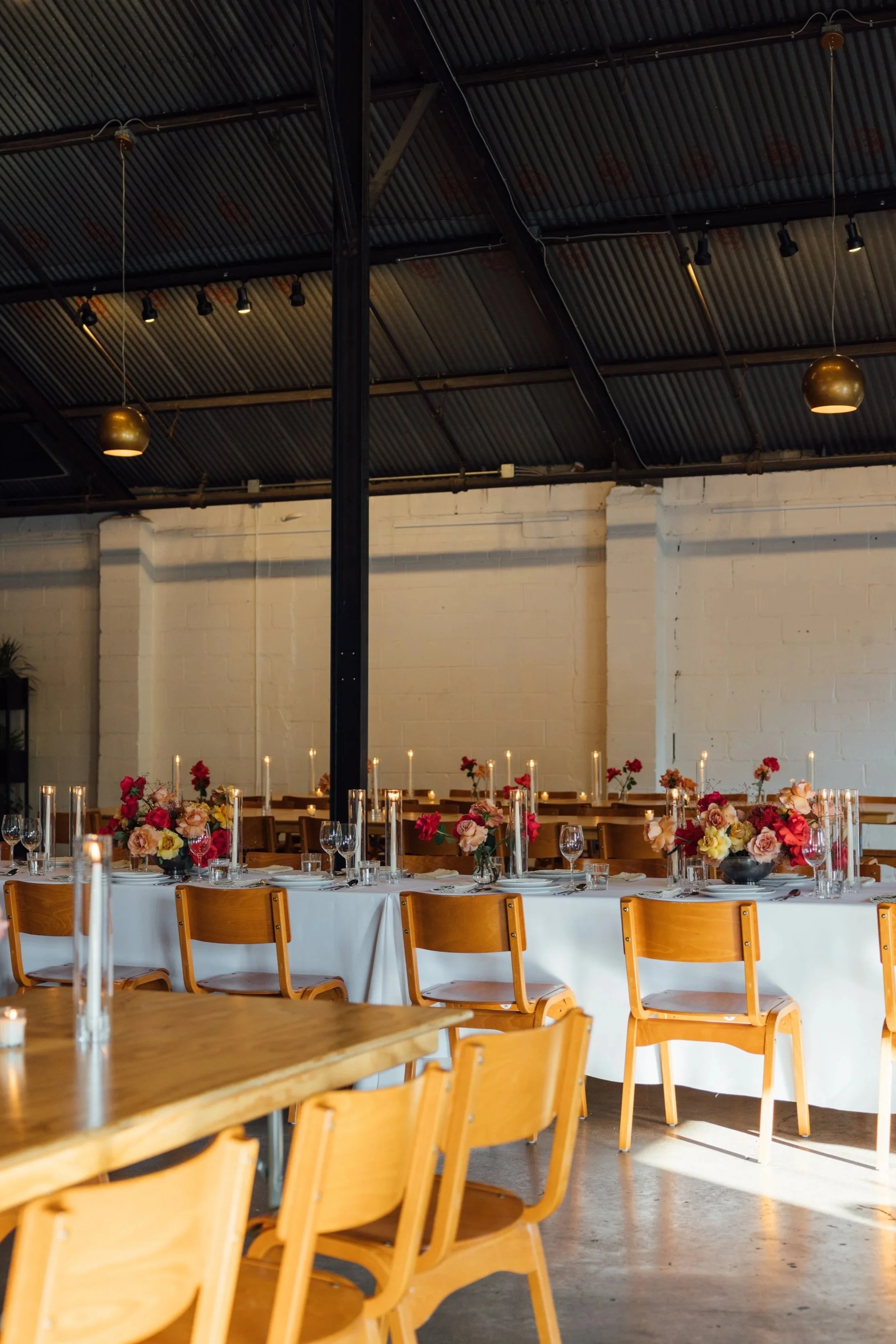 Event space with a long white table decorated with colorful flower arrangements and candles, set for a meal. Wooden chairs are arranged around the table. The room has a dark metal corrugated ceiling with hanging lights, exposed black metal beams, and a white brick wall in the background.