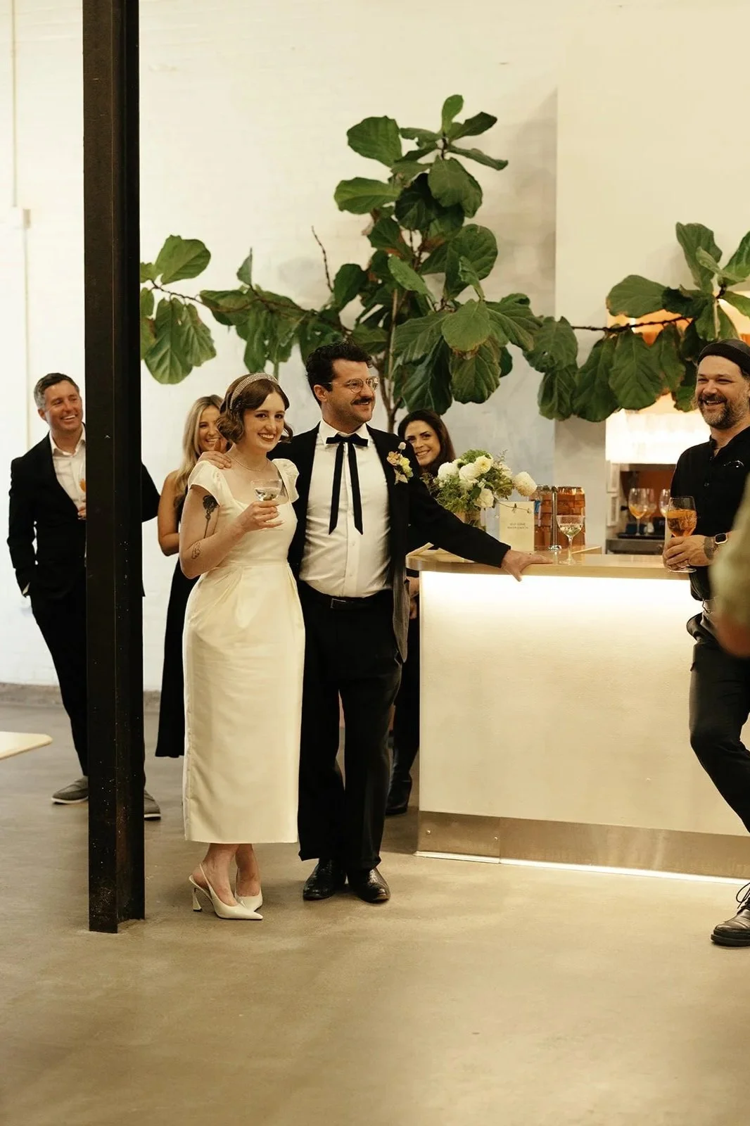 A wedding reception with a couple in wedding attire surrounded by guests. The bride wears a white dress and heels, and the groom wears a tuxedo with a bow tie. They are standing at a bar with drinks, in front of a large green plant and a white wall.