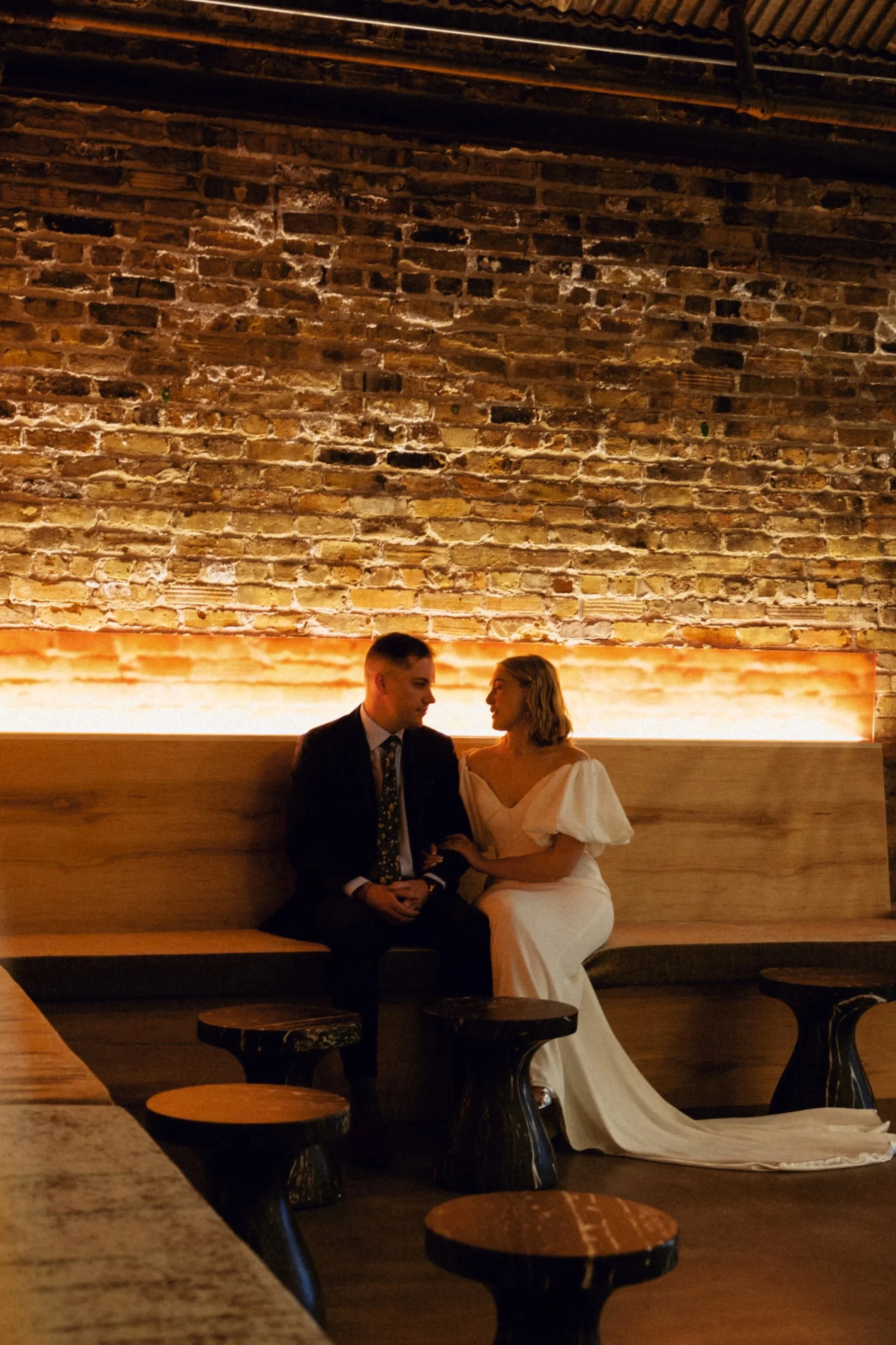 A man and woman dressed in formal attire sitting closely together on a bench in front of a brick wall with warm lighting, appearing to be having an intimate conversation near Minneapolis at a Saint Paul event venue