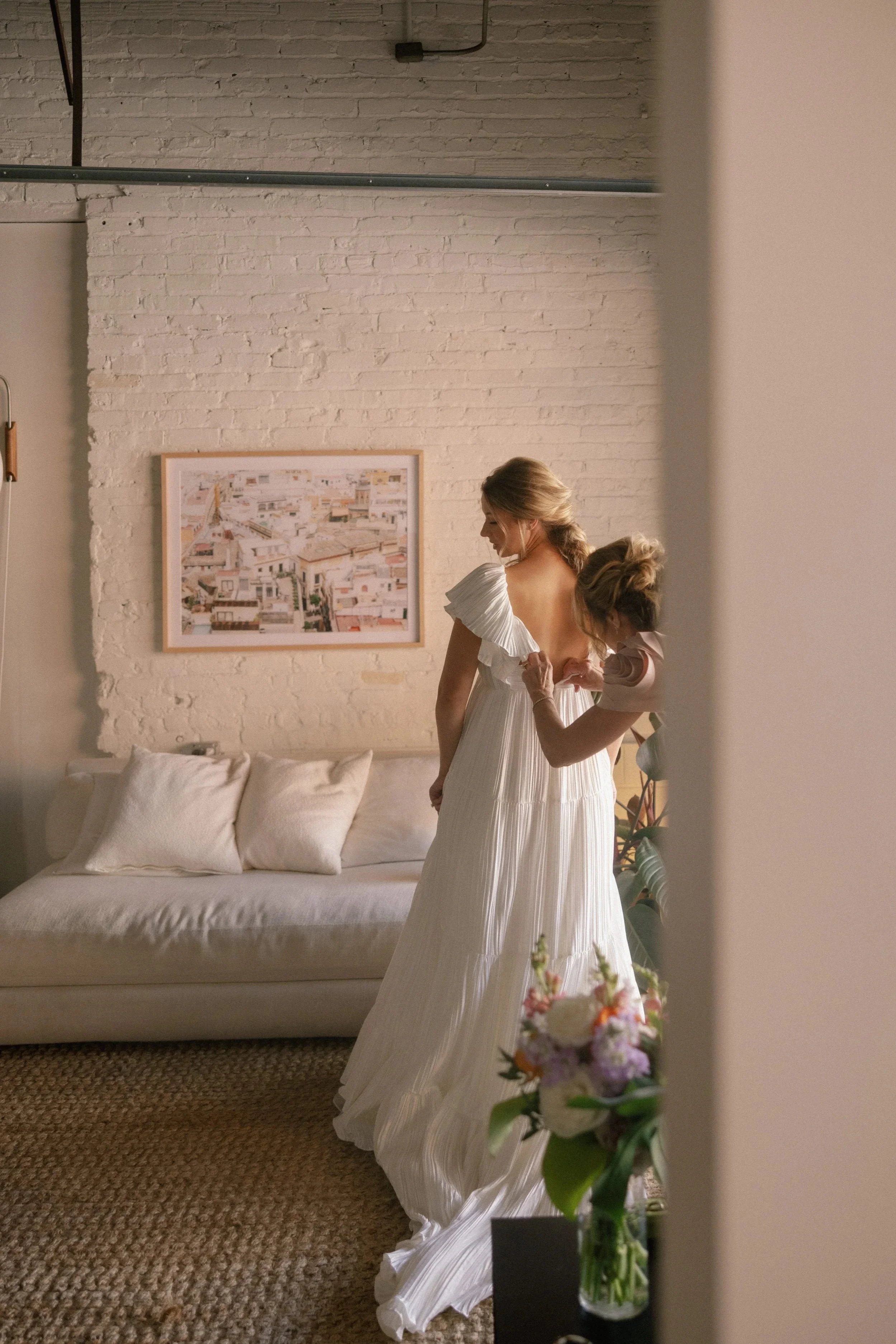 A bride getting ready in a studio or room, standing in front of a white-painted brick wall with a framed cityscape photograph, wearing a white dress, with an assistant helping her with the back of her dress, near a white sofa with pillows, with a vase of flowers in the foreground.
