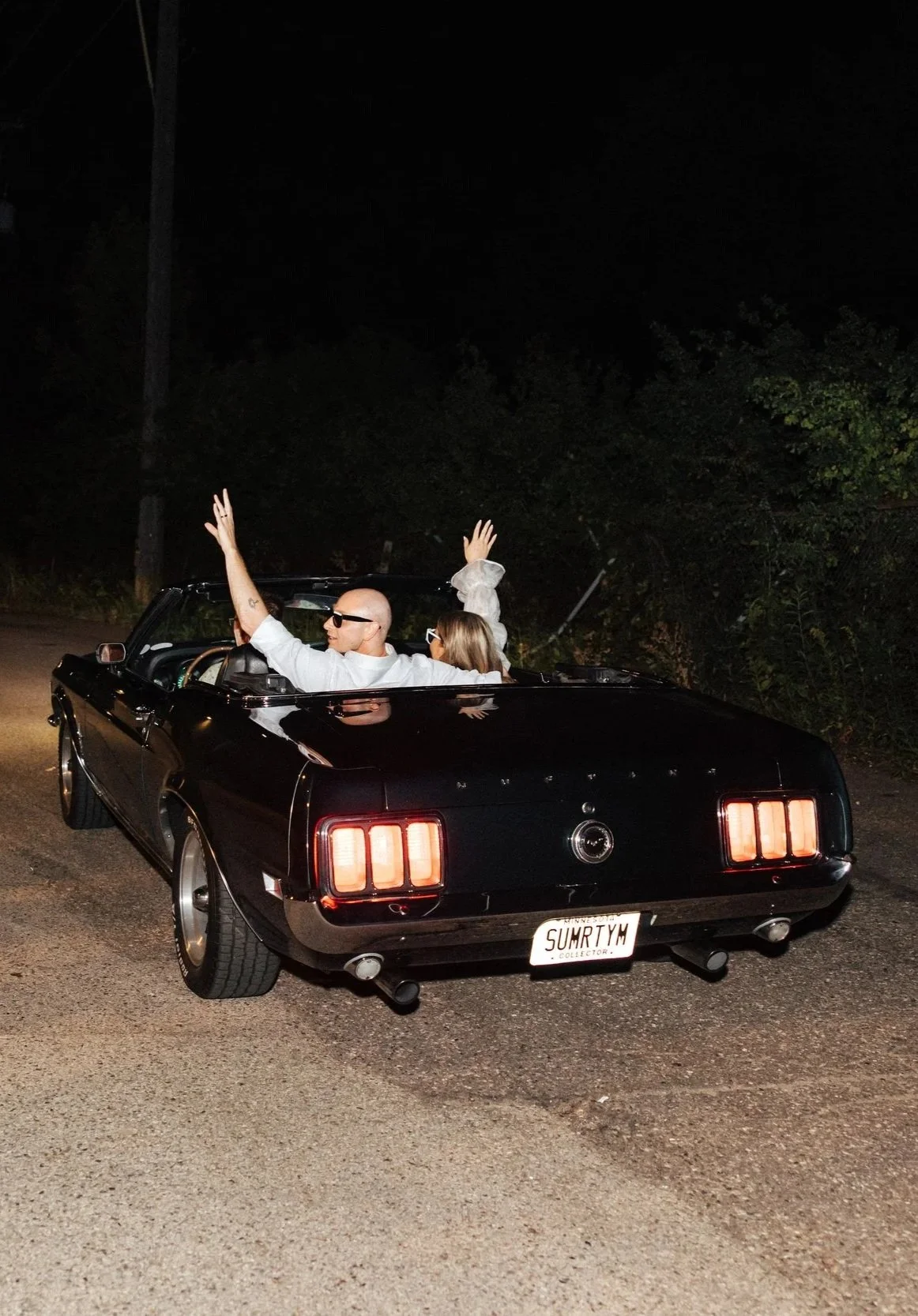 A black vintage convertible car with a man and woman inside at night, the man is bald, wearing sunglasses, and waving, while the woman has shoulder-length hair and is wearing sunglasses - a wedding couple depart from their Saint Paul wedding.