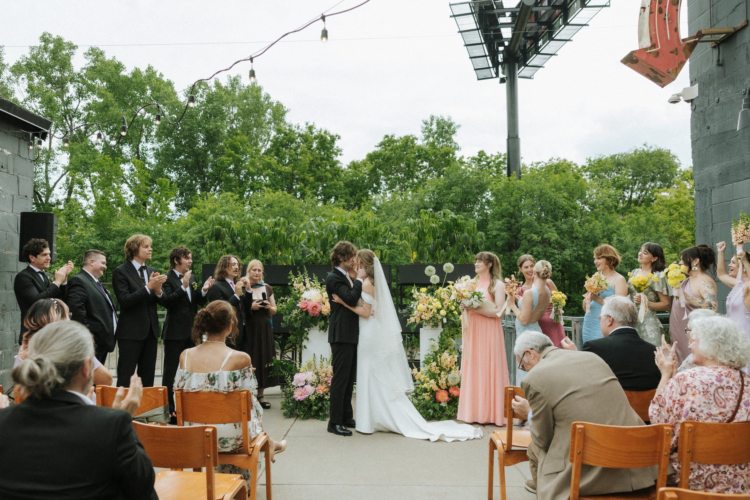 A wedding ceremony taking place outdoors with the bride and groom kissing, surrounded by wedding guests seated in chairs and standing, with trees and overhead string lights in the background at PAIKKA, a St. Paul wedding venue.