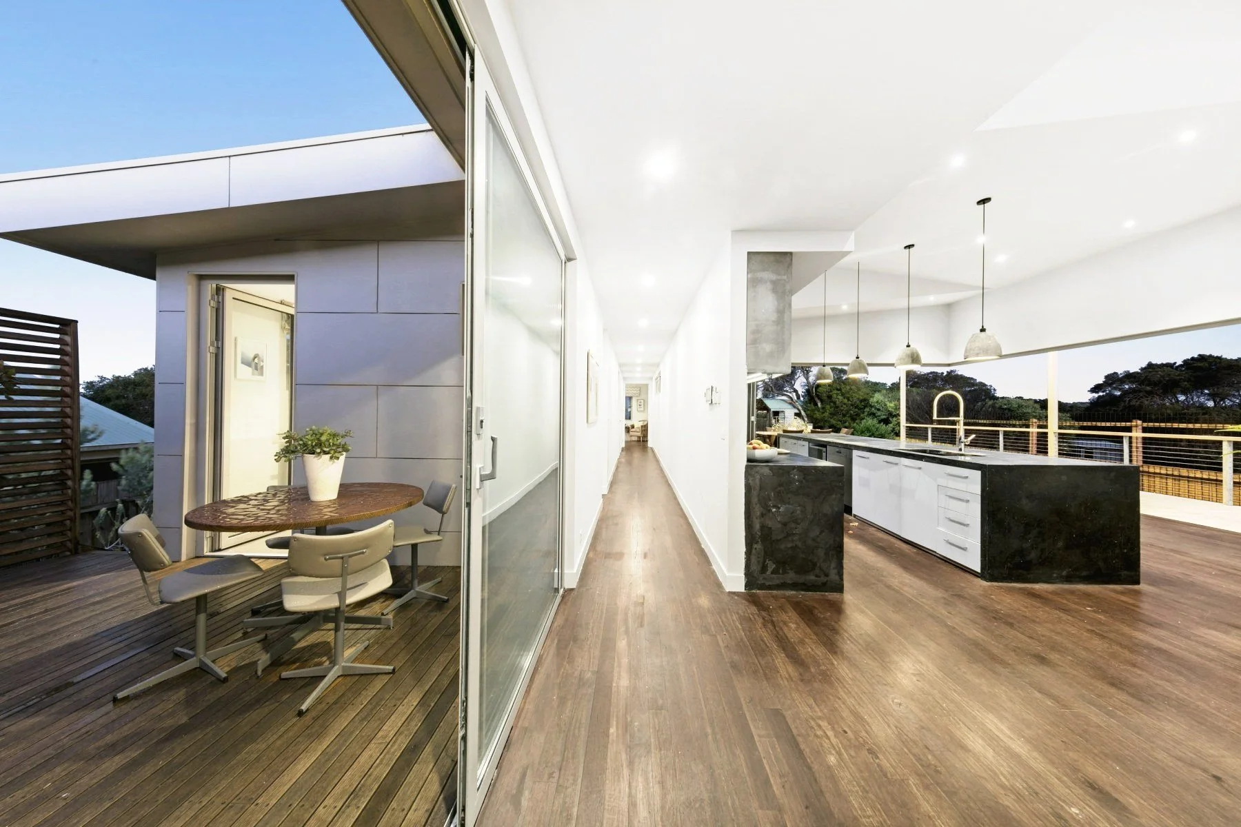 Modern outdoor balcony and interior kitchen of a house with wooden floors and white walls, featuring black and white kitchen island, hanging pendant lights, and outdoor seating area with a round table and chairs.