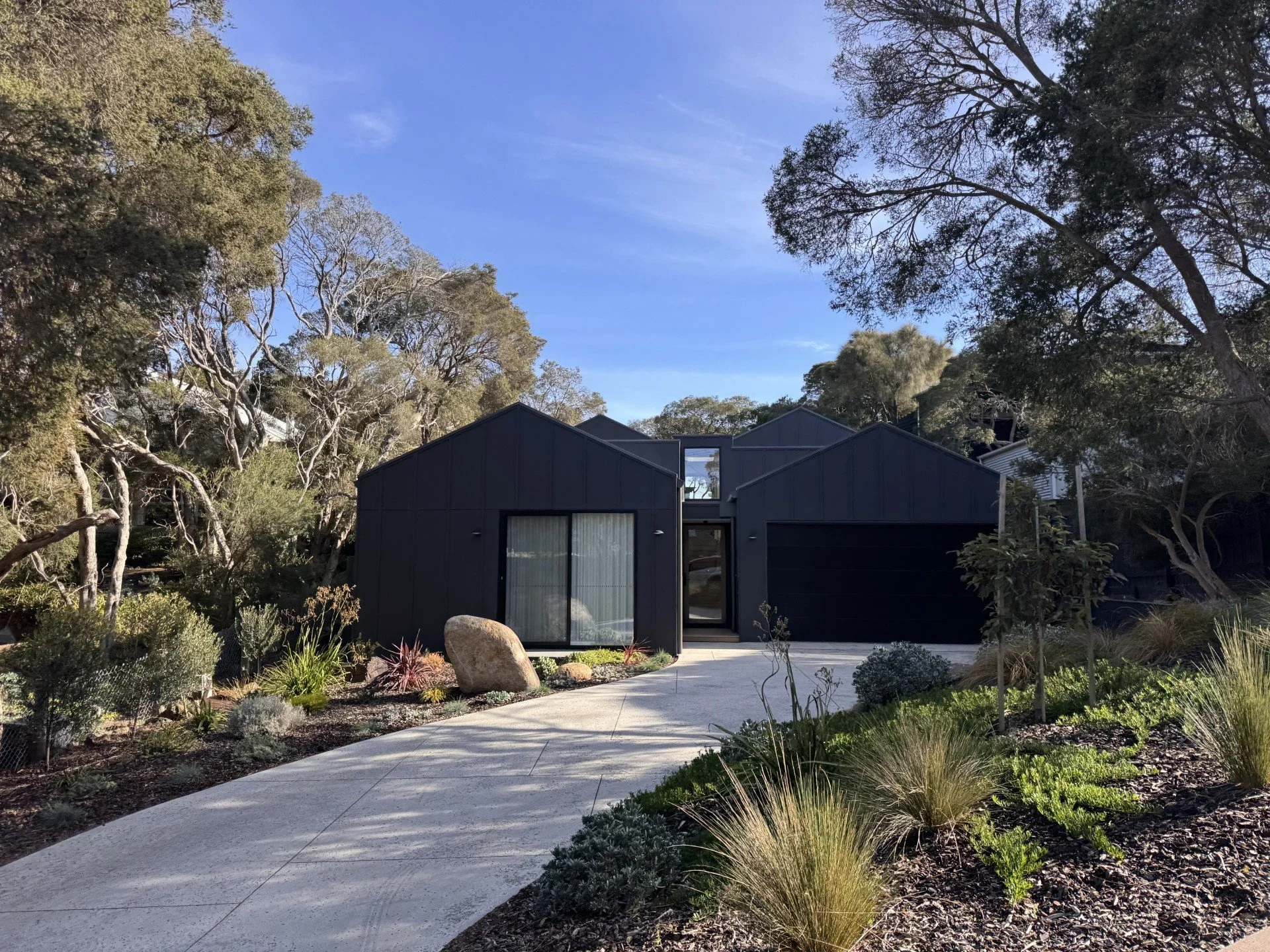 Modern dark gray house with large windows and a surrounding garden, viewed from a curved concrete pathway amidst trees and bushes under a clear blue sky.