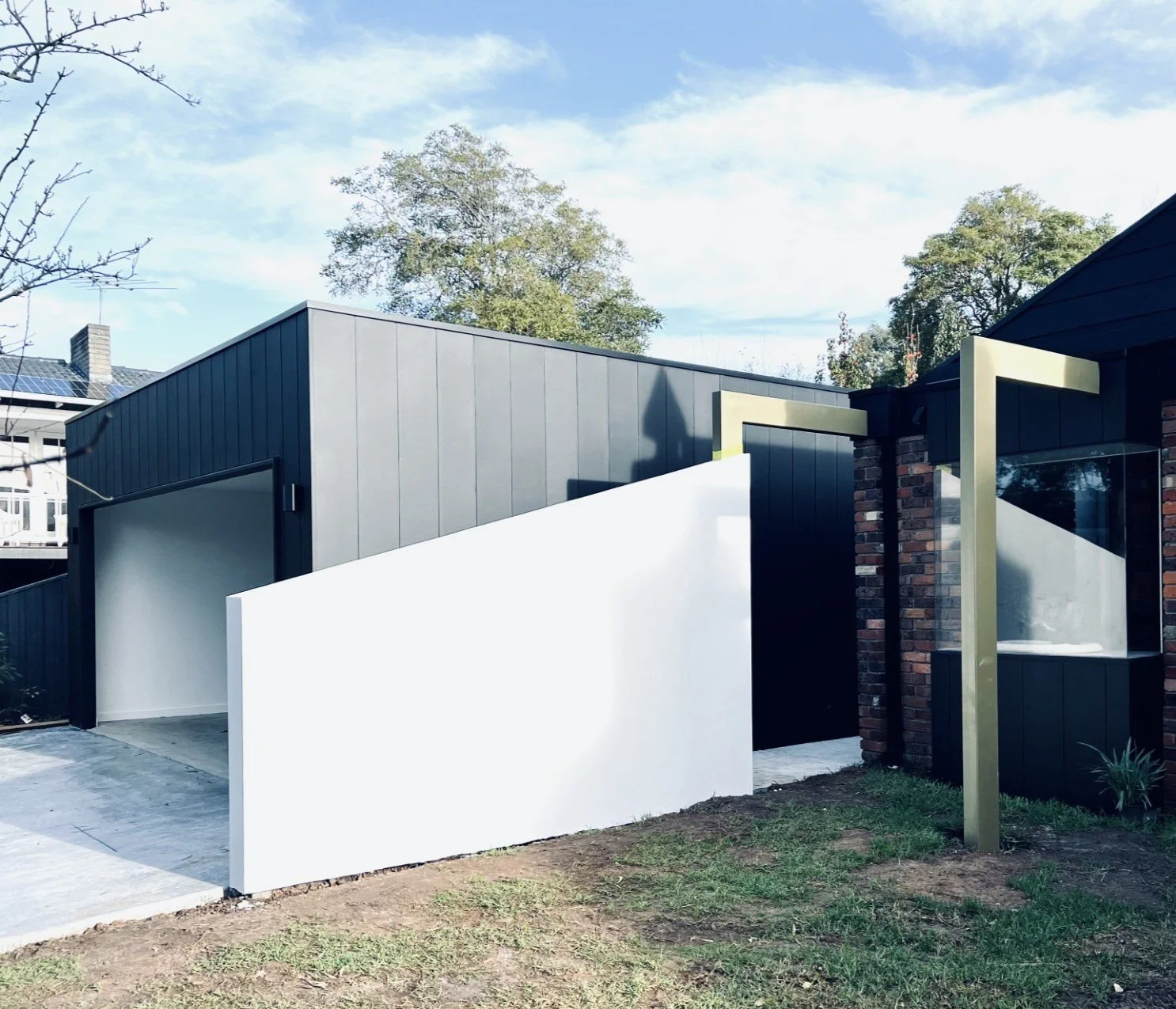 Modern house with black and gray exterior, gold accents, brick wall, large window, white fence, and a tree in a yard under a partly cloudy sky.