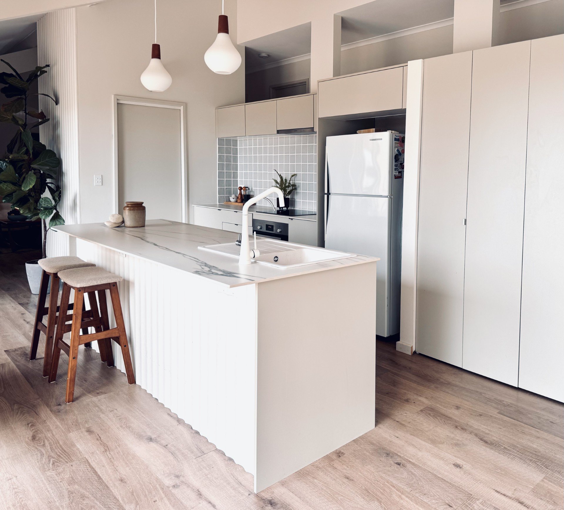 Modern kitchen with white cabinetry, marble kitchen island, and wooden bar stools. There are pendant lights overhead, a refrigerator, a plant, and decorative jars on the counter. The floor is wood, and the walls are painted white.