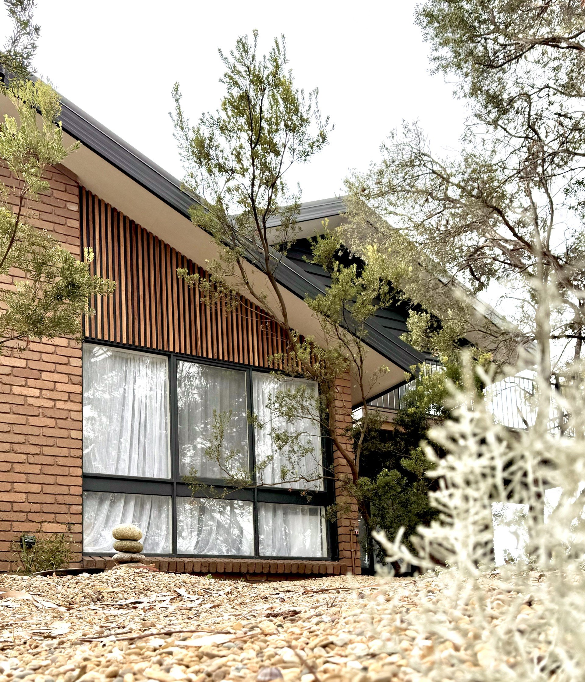 Modern house with brick and wood exterior, large corner window, surrounded by trees and a gravel yard with a small stone sculpture.