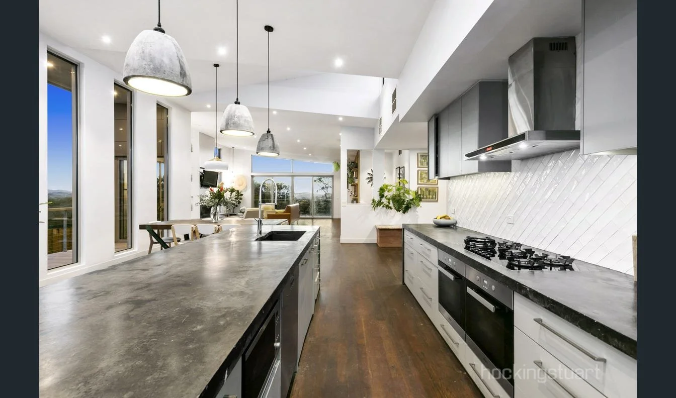 Modern kitchen with dark countertops, white cabinets, stainless steel appliances, and concrete pendant lights, opening to a living area with large windows and outdoor view.