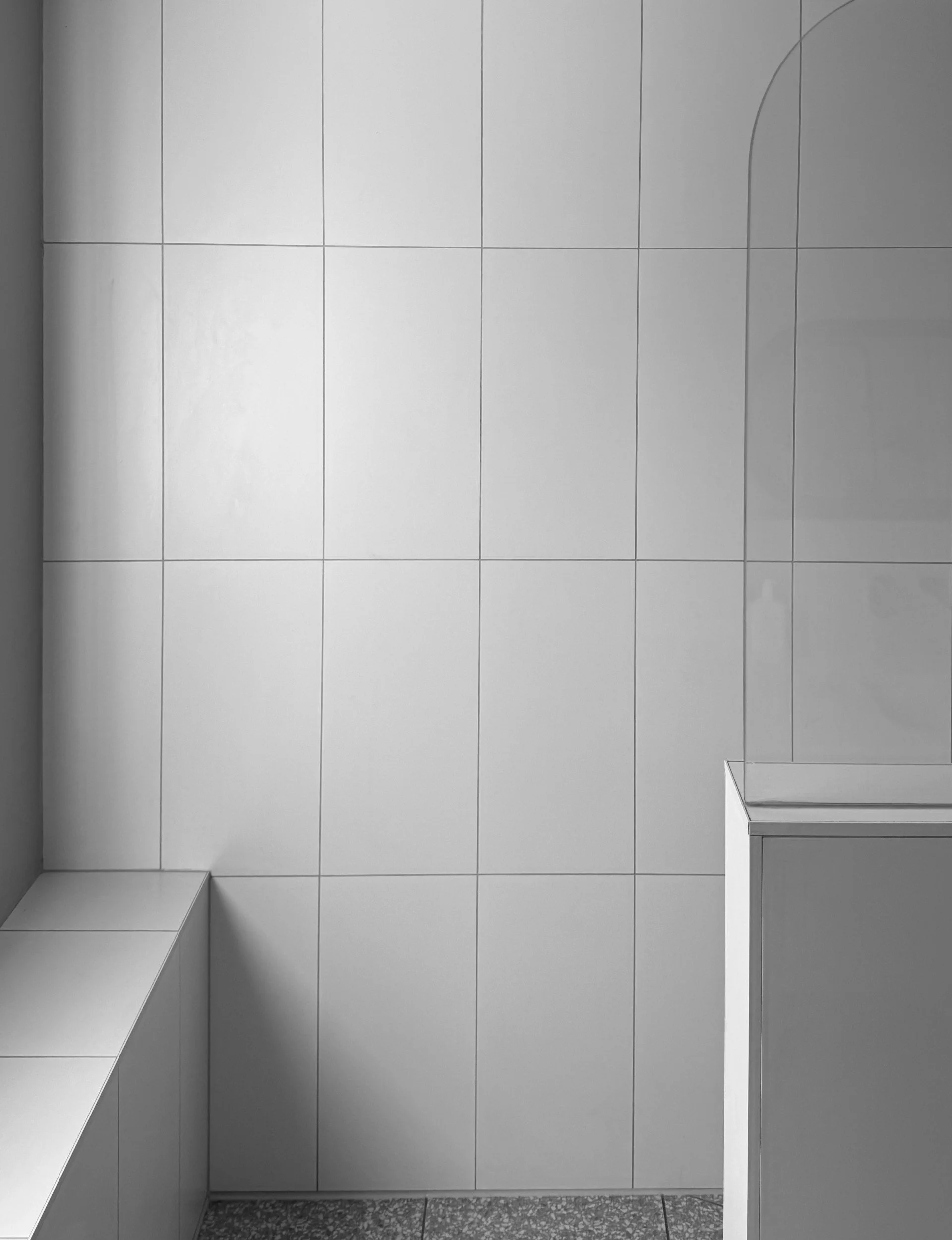 A small bathroom with white tiled walls and a textured gray tile floor. There is a built-in shelf on the left wall and a partial view of a mirror or panel on the right side.