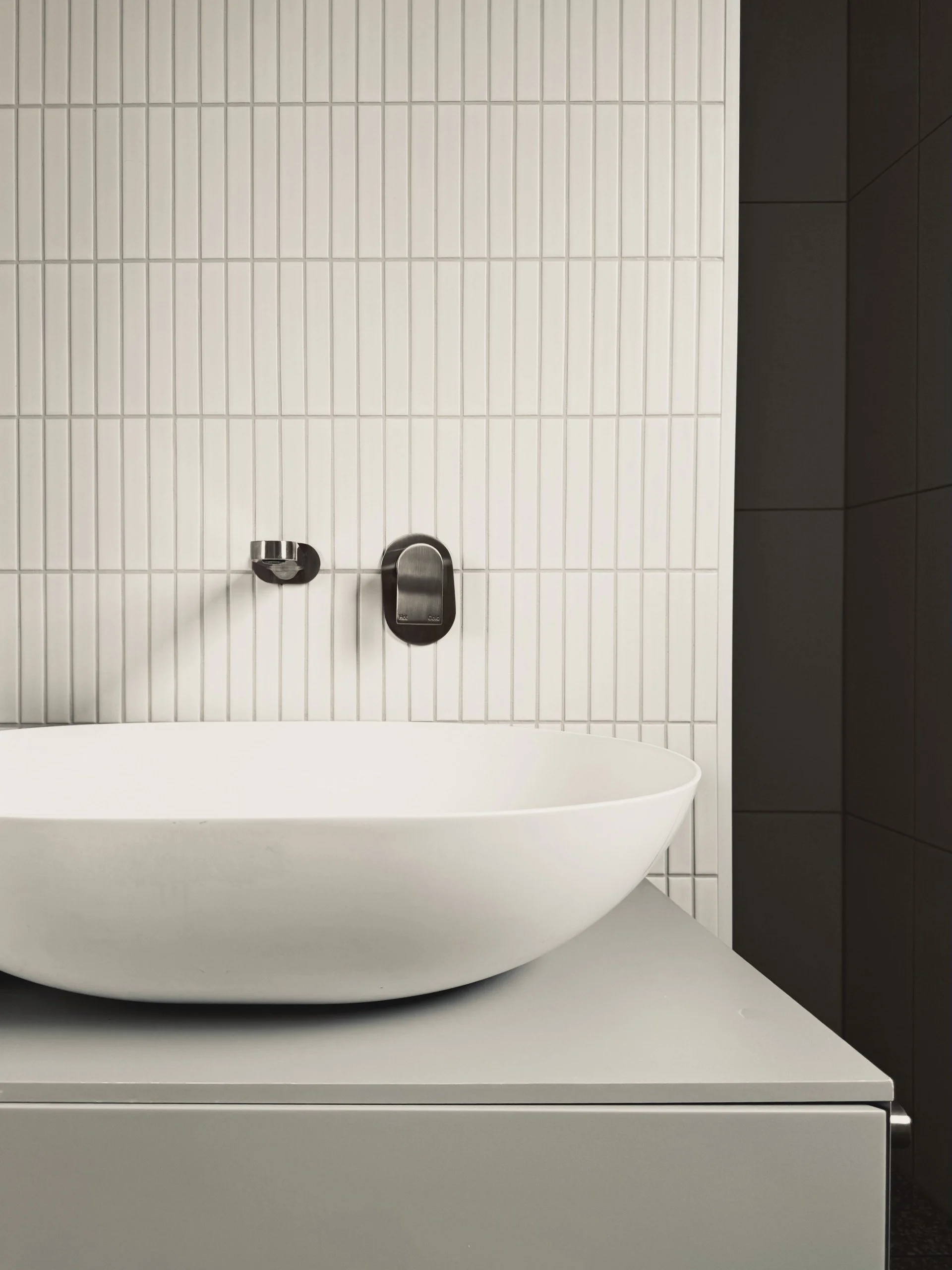 A minimalist bathroom with white tiled wall, a white oval bathtub, a silver faucet, and dark cabinetry on the right.