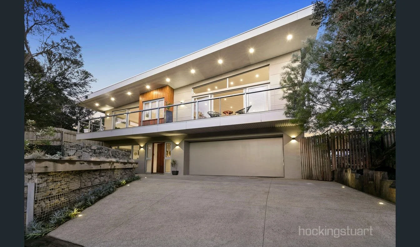 Modern two-story house with a flat roof, outdoor balcony, and large windows, illuminated by exterior lights at dusk.