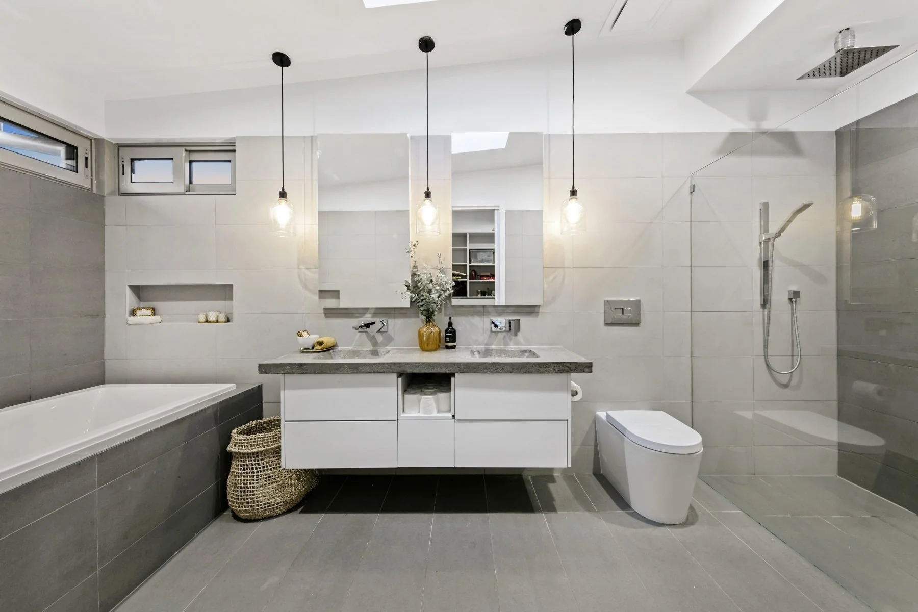 Modern bathroom with gray tiles, white walls, a built-in bathtub, a floating vanity with a gray countertop, a mirror, pendant lights, a yellow vase with flowers, a toilet, and a walk-in shower with a rainfall showerhead.
