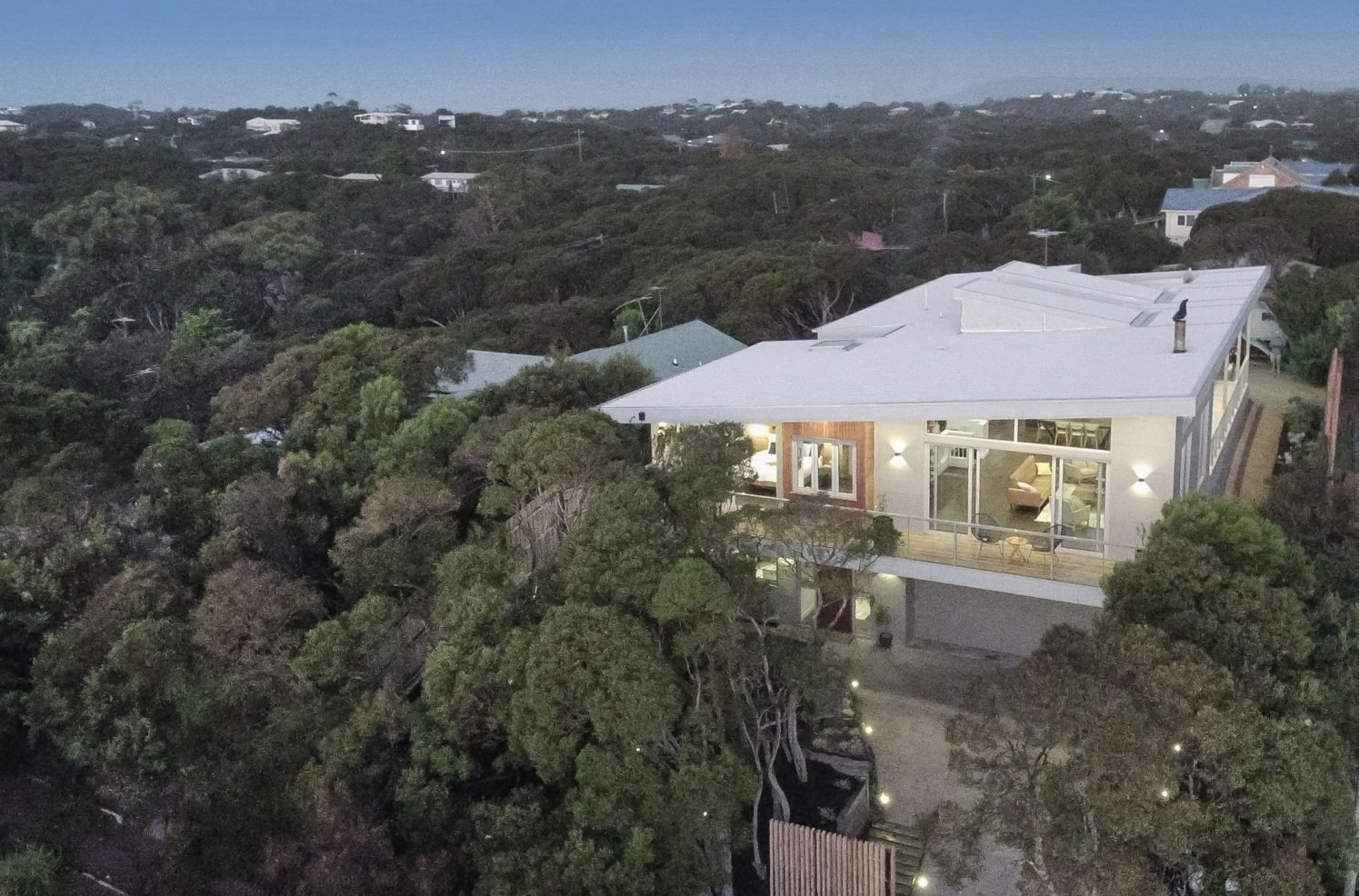 Aerial view of a modern house with large glass windows, surrounded by trees in a suburban neighborhood during twilight.