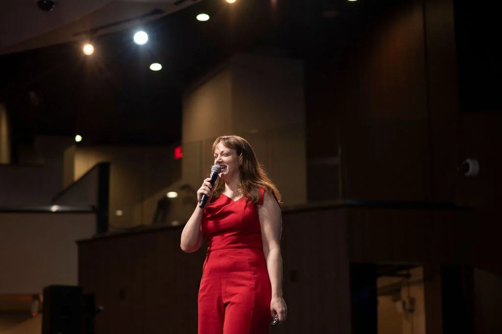 Nina Nesdoly smiling and speaking on stage in red outfit, holding microphone and smiling
