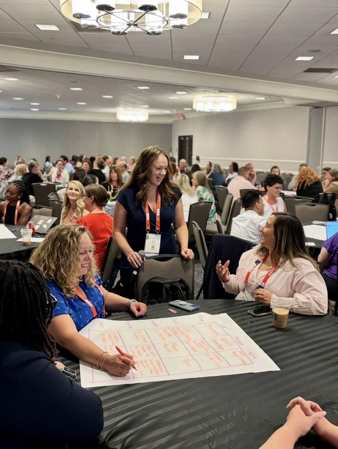 Two women at a conference table speaking to the facilitator during a workshop with other conference tables and people in the background.