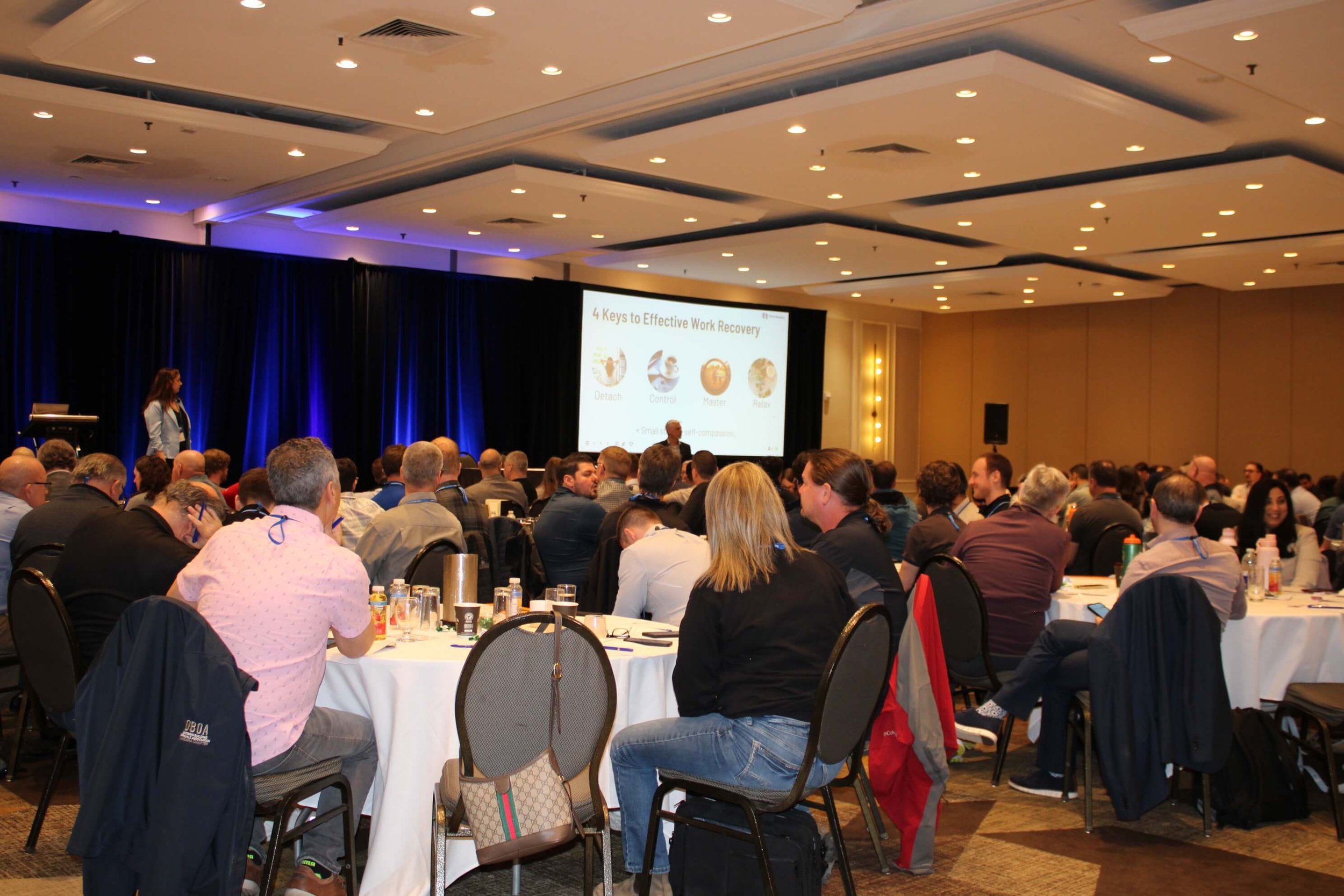 Conference audience seated at round tables watching Nina Nesdoly present keynote on work recovery and burnout prevention