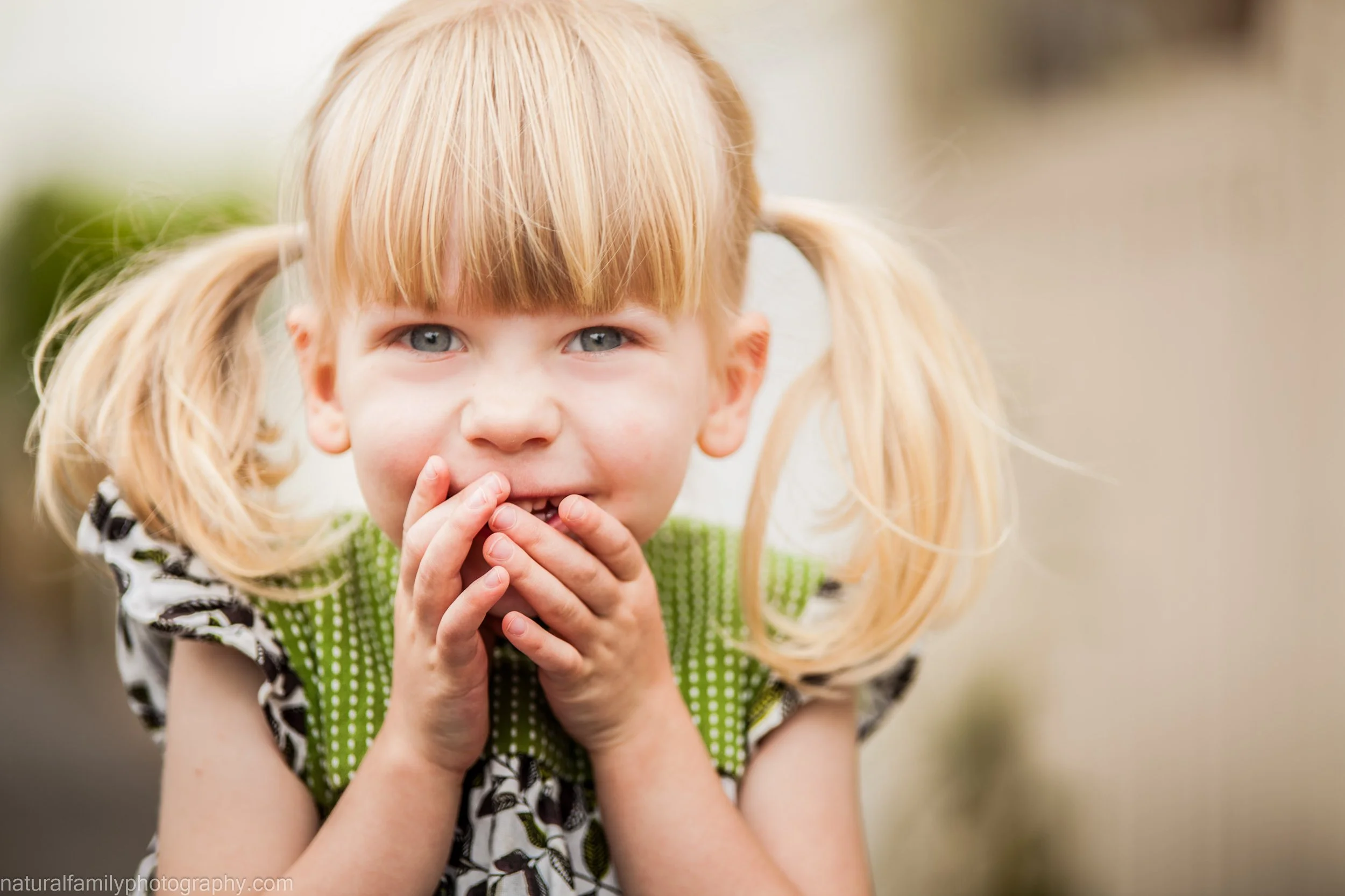 A young girl with blonde hair in pigtails and wide blue eyes, wearing a green polka dot top, holding her hands over her mouth with a playful or surprised expression. Portrait by Natural Family Photography, Melbourne.