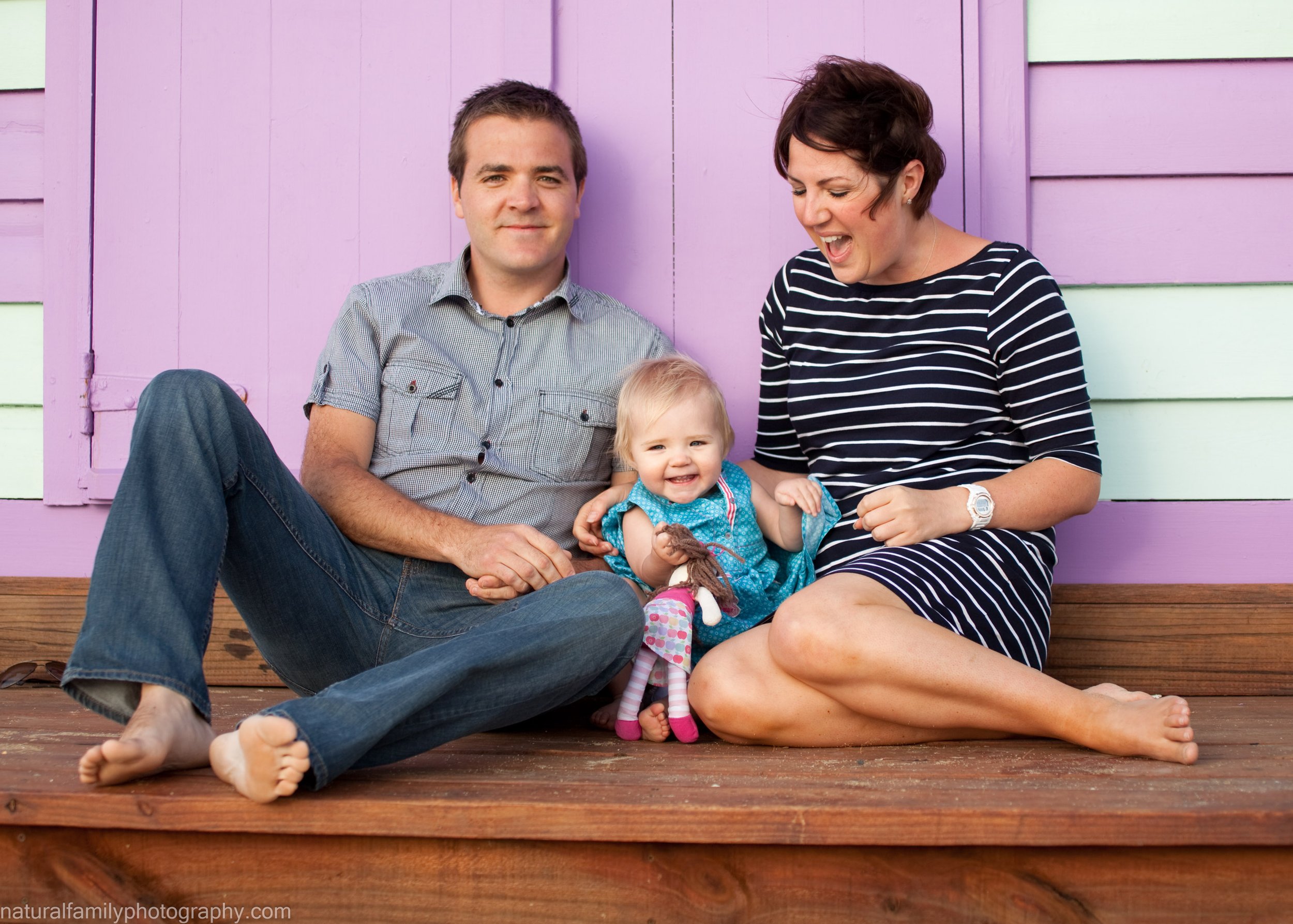 Family of three (father, mother, and toddler girl) sitting on a wooden porch in front of a pink and white beach box in Brighton, smiling and playing together. Portrait by Natural Family Photography, Melbourne.