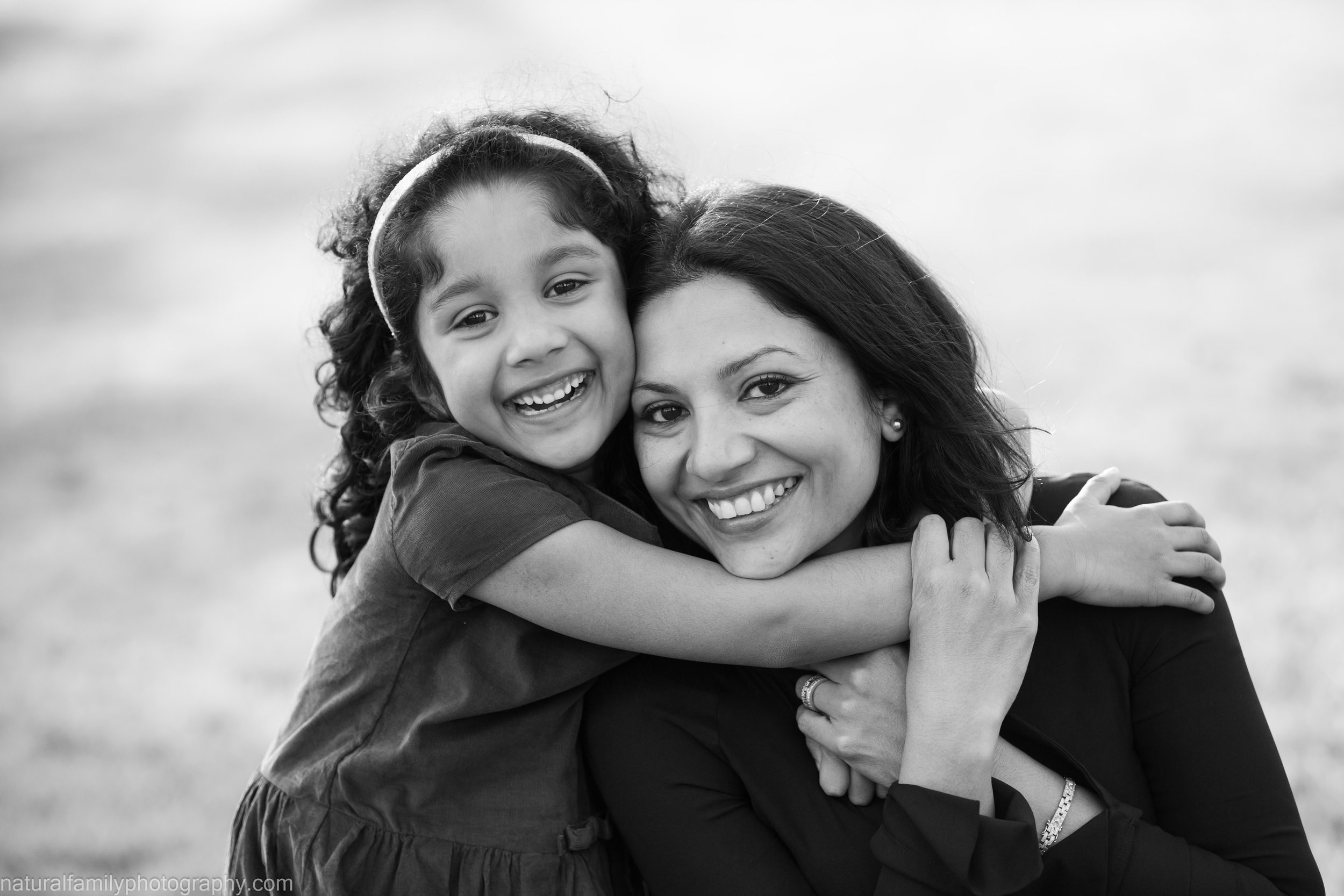 A smiling woman holding a young girl in an embrace outdoors with a blurred background.