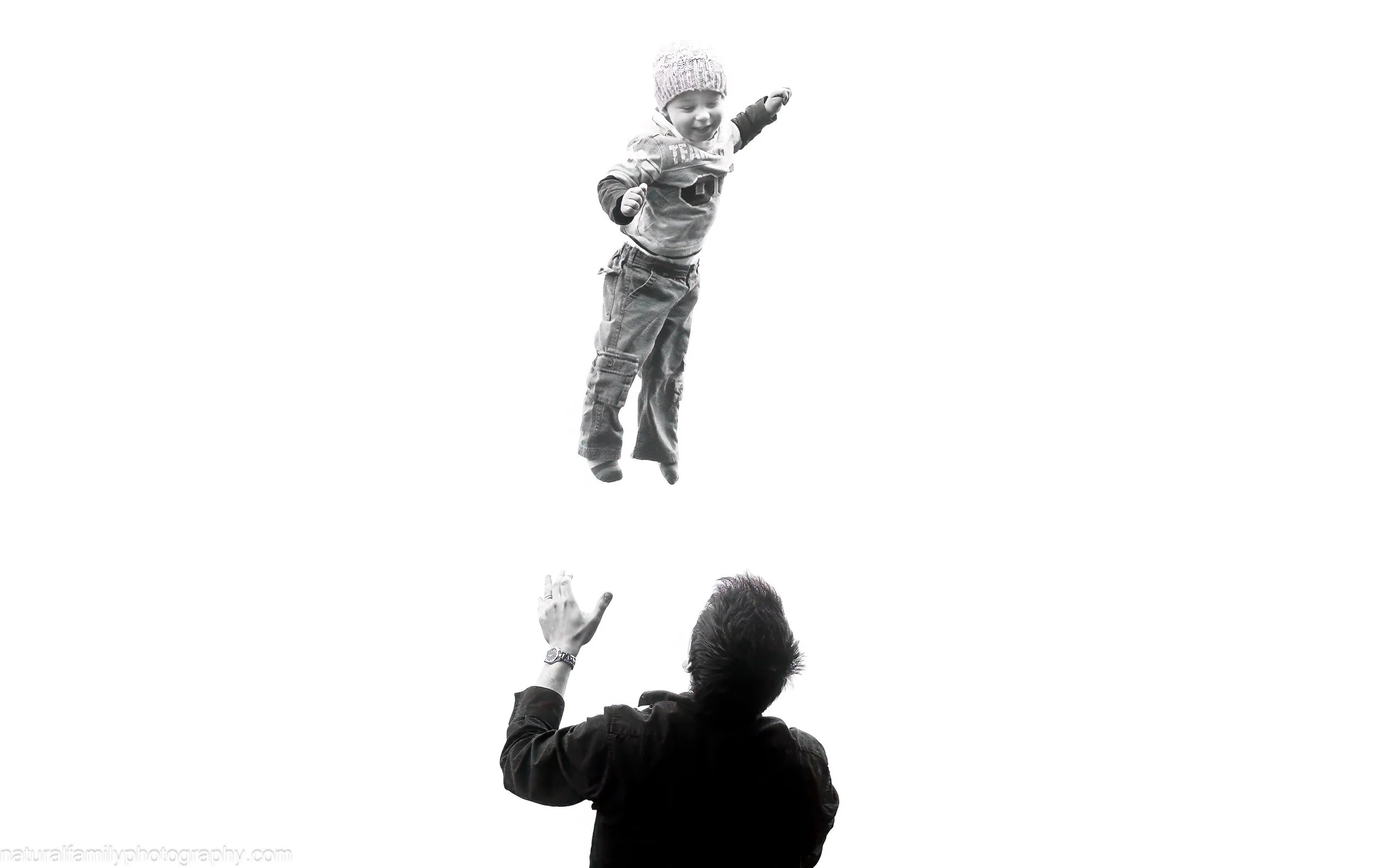 A black and white photo of a young boy being thrown in the air by his father, underneath him. Action portrait by Natural Family Photography, Melbourne.