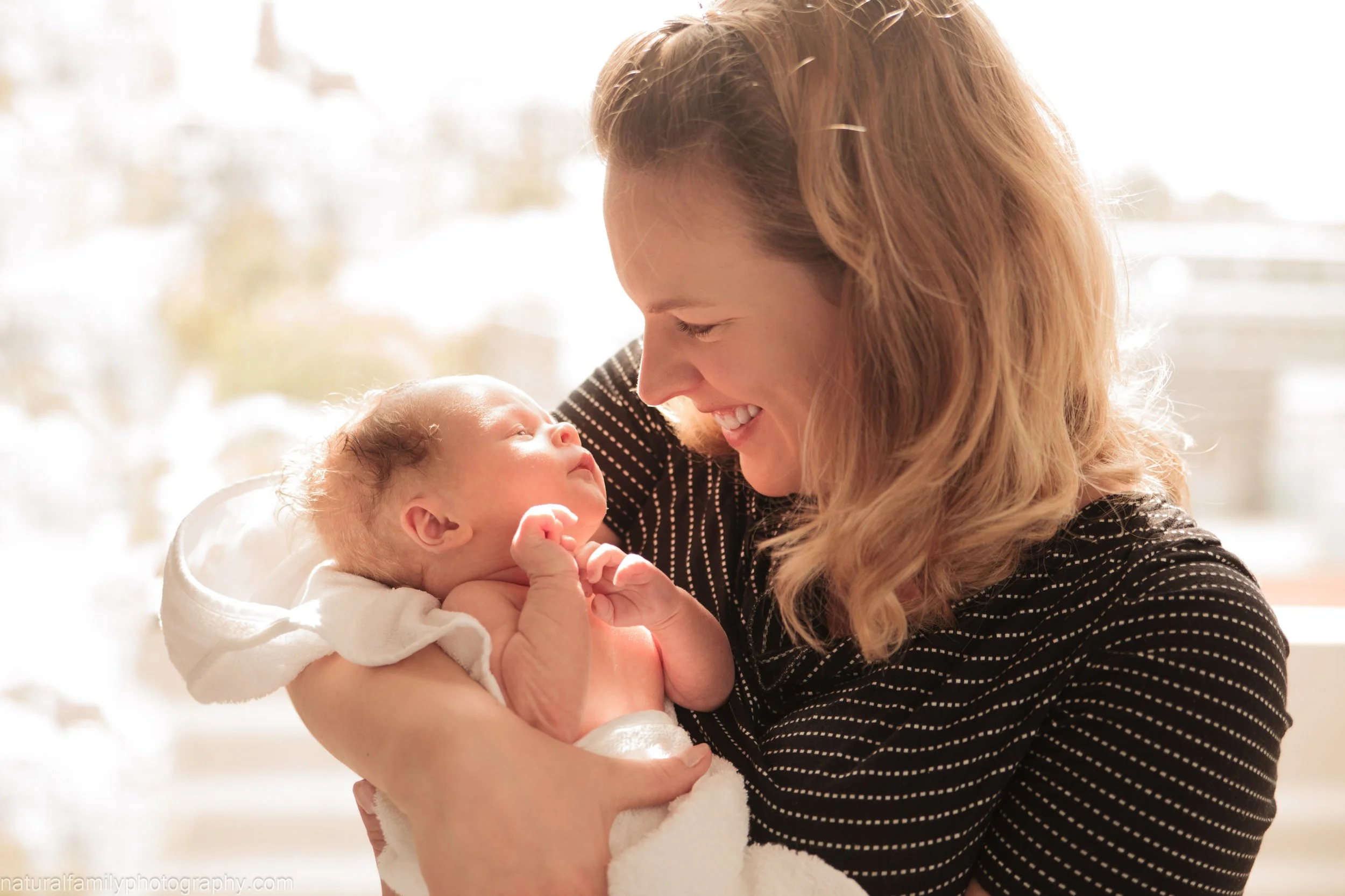 A woman holding a newborn baby close, both smiling and looking at each other indoors with bright natural light in the background.