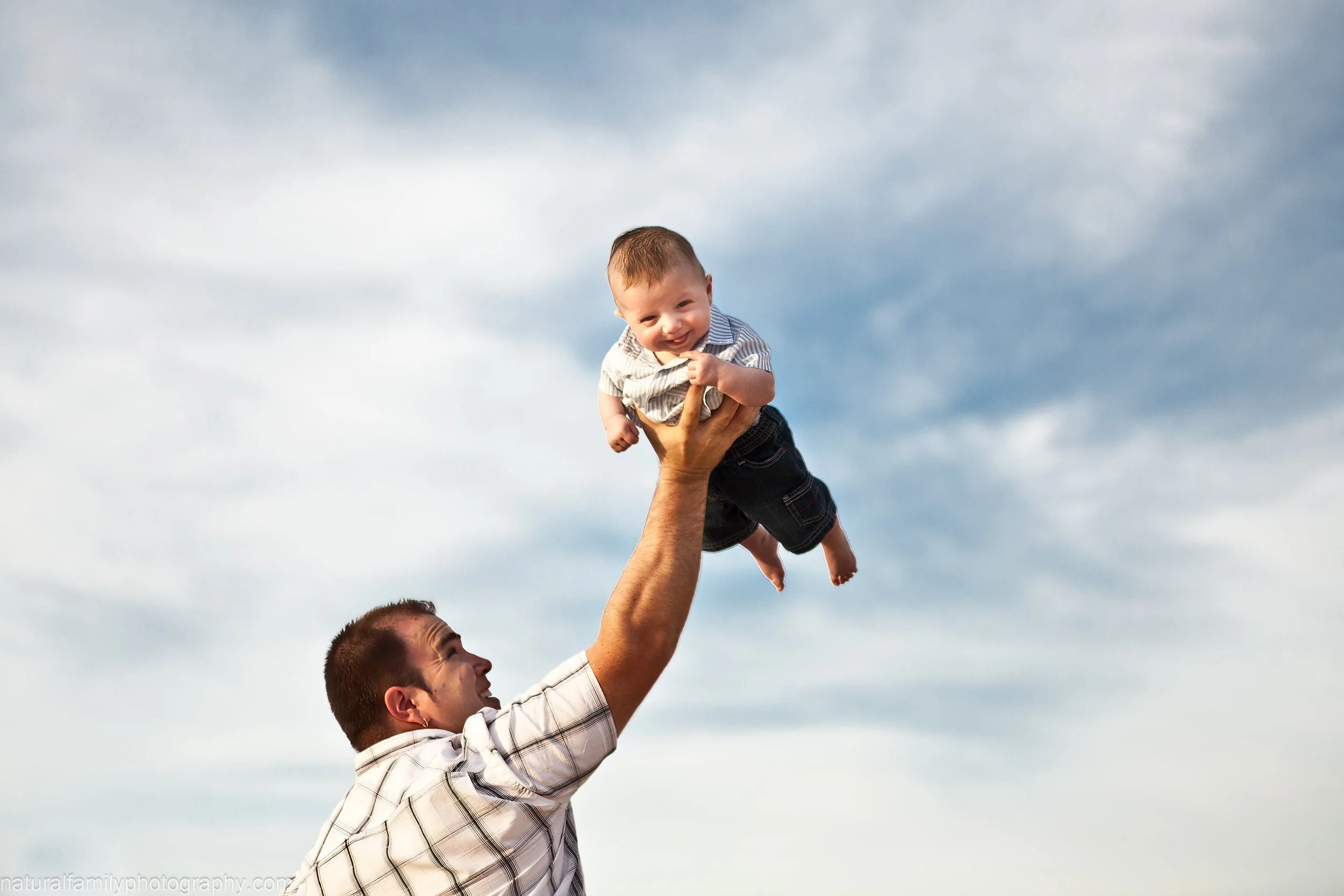 A man lifts a smiling baby into the air outdoors against a cloudy sky with one hand. Joyful portrait by Natural Family Photography, Melbourne.