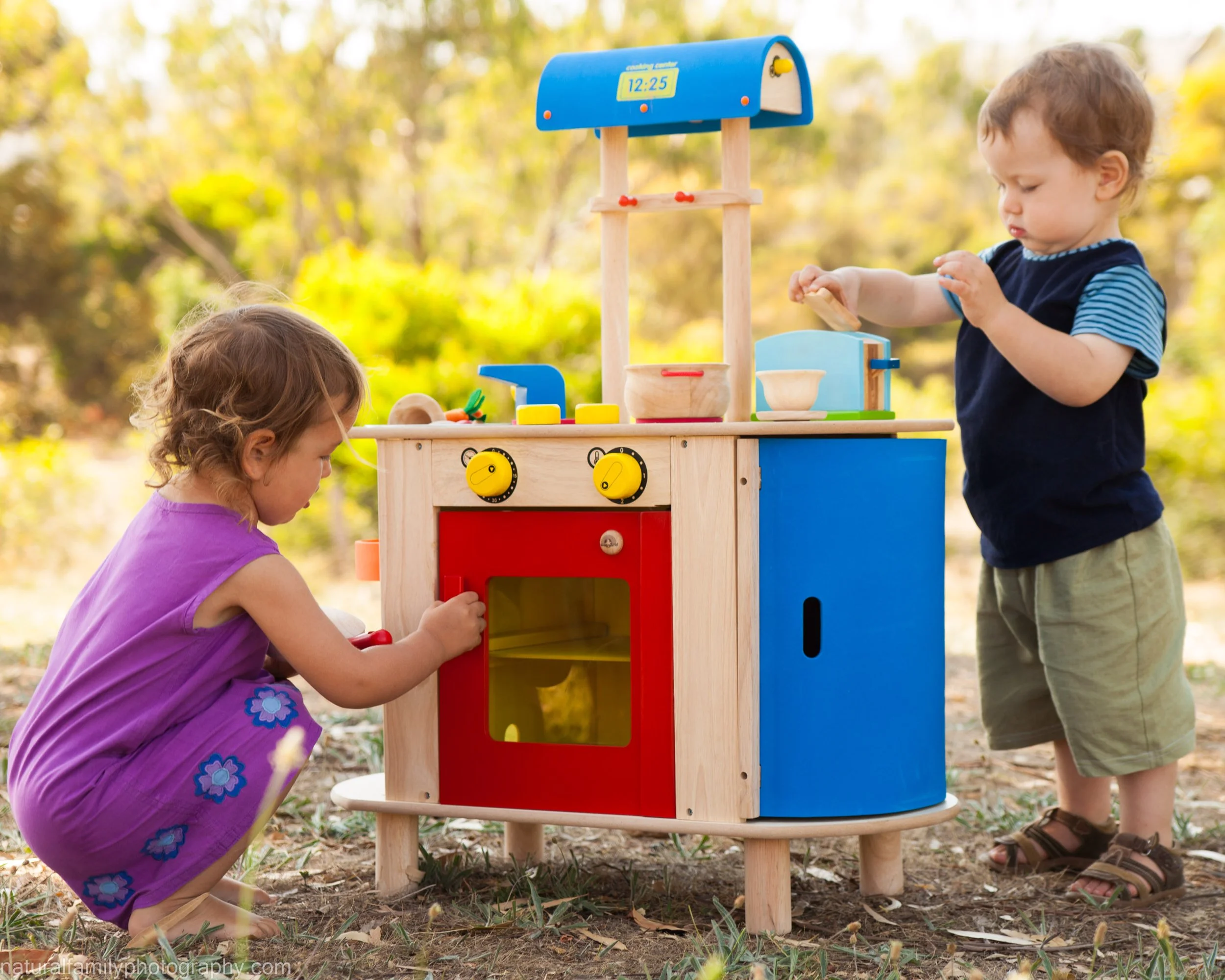 Two young children playing with a wooden toy kitchen outdoors, surrounded by trees and grass.