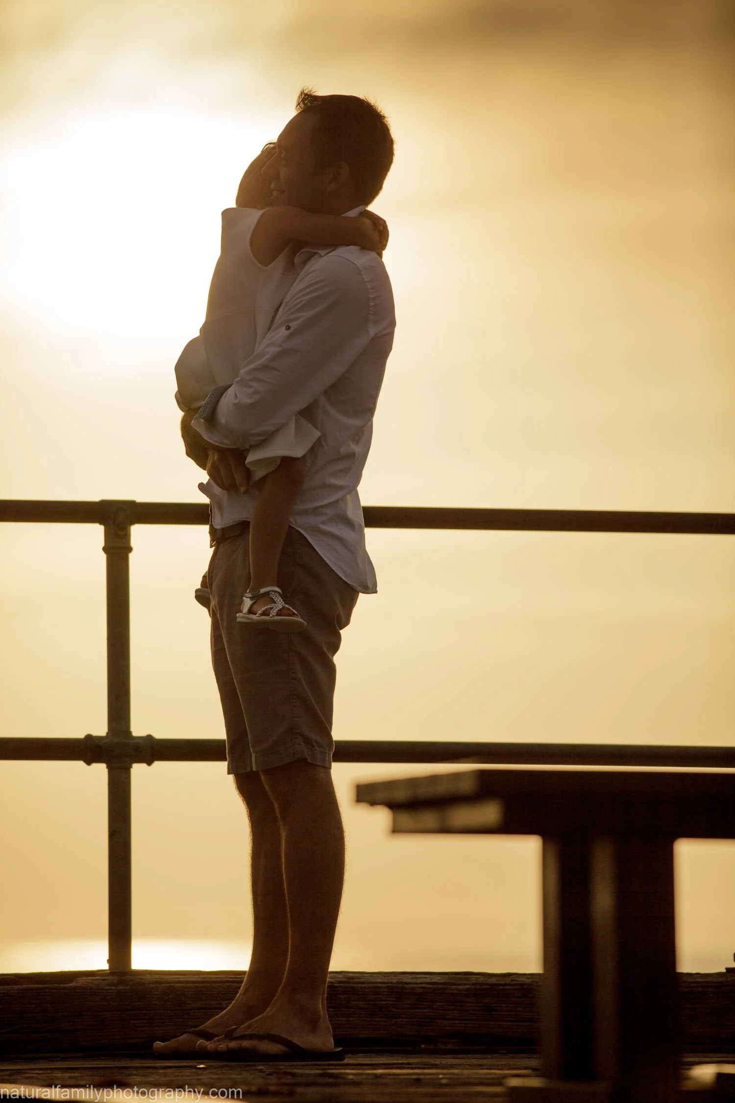 A man holding a child in his arms, silhouetted against a sunset or sunrise, standing on a platform with a railing.