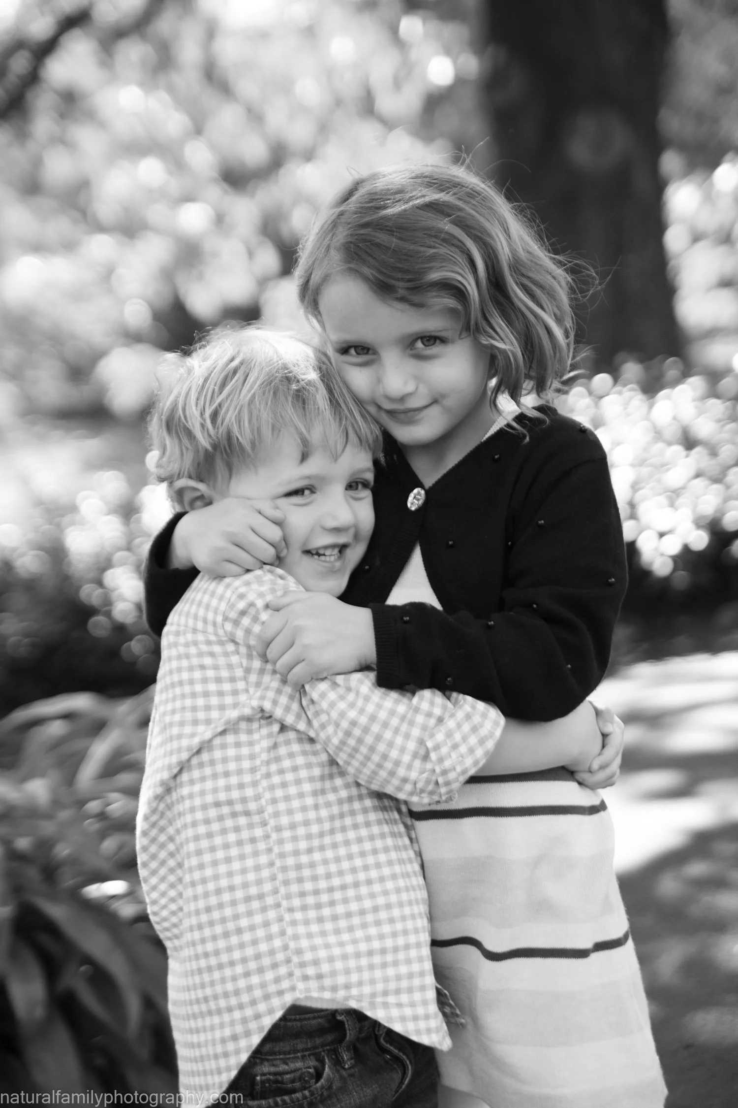 Two children, a boy and a girl, hugging and smiling outdoors with trees in the background.
