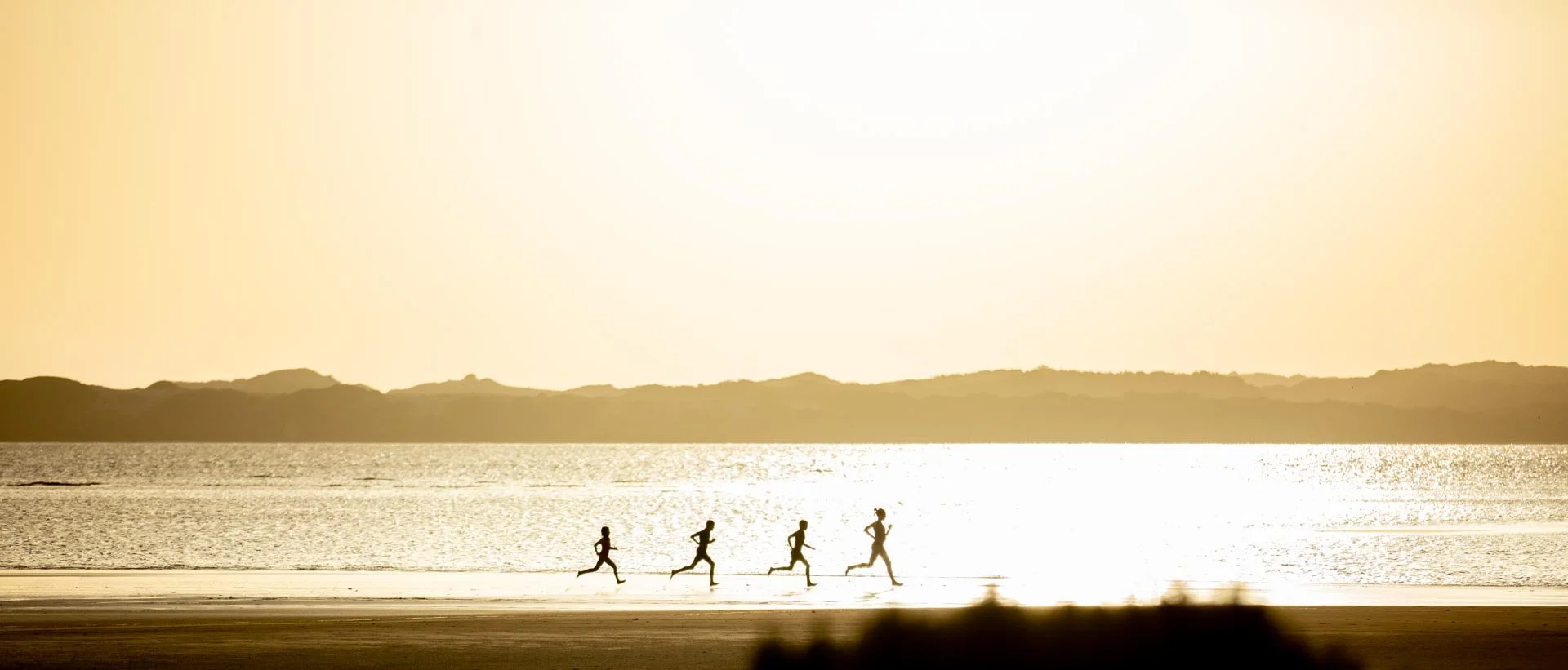 Four people jogging along a beach at sunset with the ocean in the background